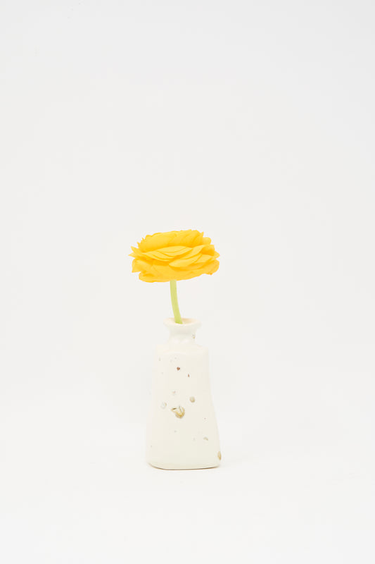A single yellow flower with a green stem sits in the small, white Carved Bud Vase I by MONDAYS against a plain white background.