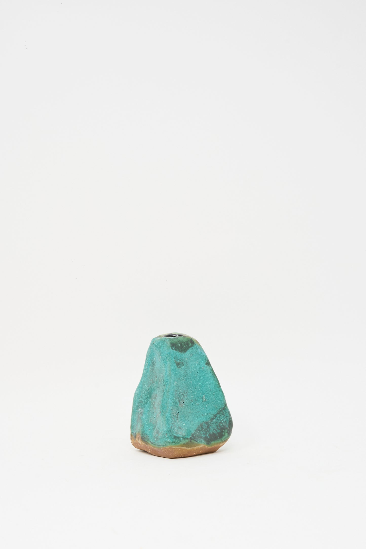 The Carved Bud Vase III by MONDAYS is a small, irregularly shaped clay vase with a textured turquoise surface and a brown base, shown against a plain white background.