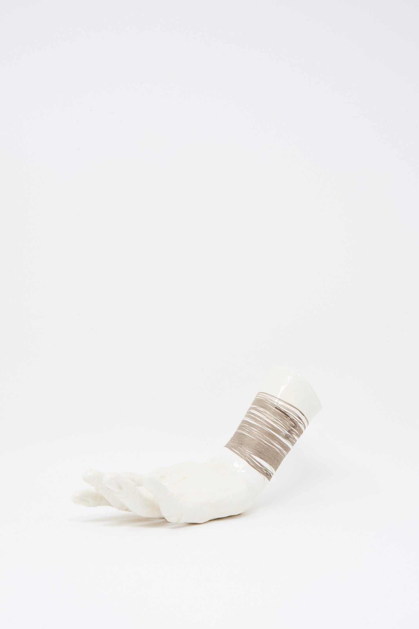 The Ceramic Hand Sculpture with Grey Thread by Monty J features a white ceramic hand lying on its side, with the wrist wrapped in beige and grey thread. Handmade in Brooklyn.