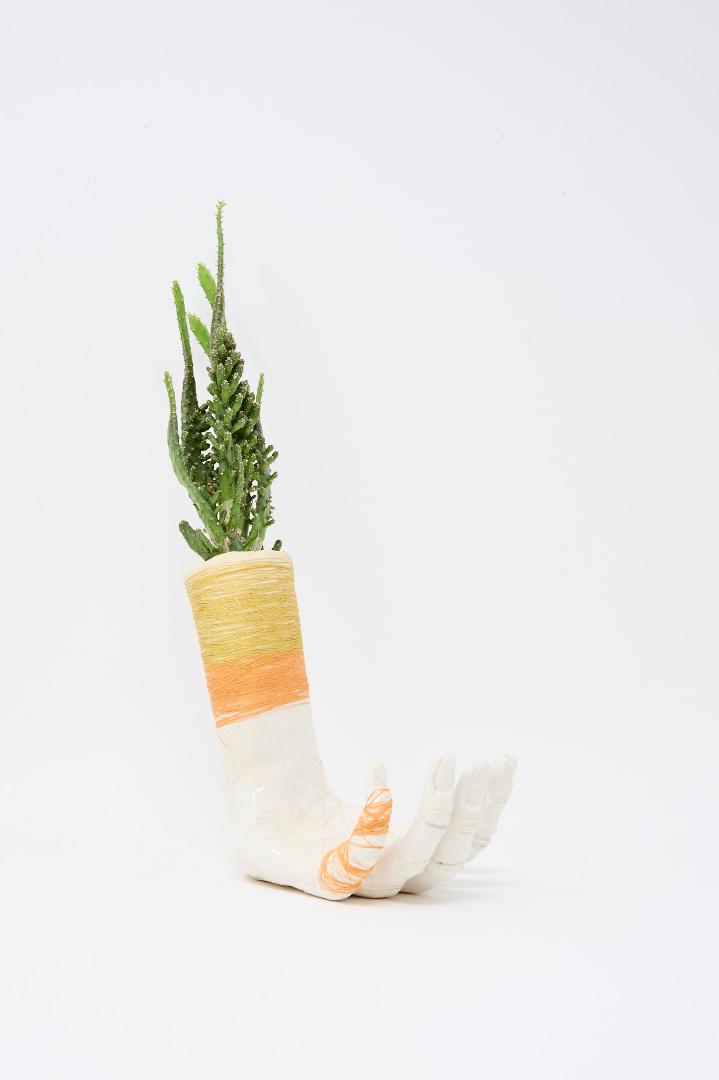 The Monty J Ceramic Hand Sculpture with Yellow and Orange Thread, handmade in Brooklyn, is designed to hold green succulents or cacti, featuring vibrant threads wrapped around the wrist and fingers.