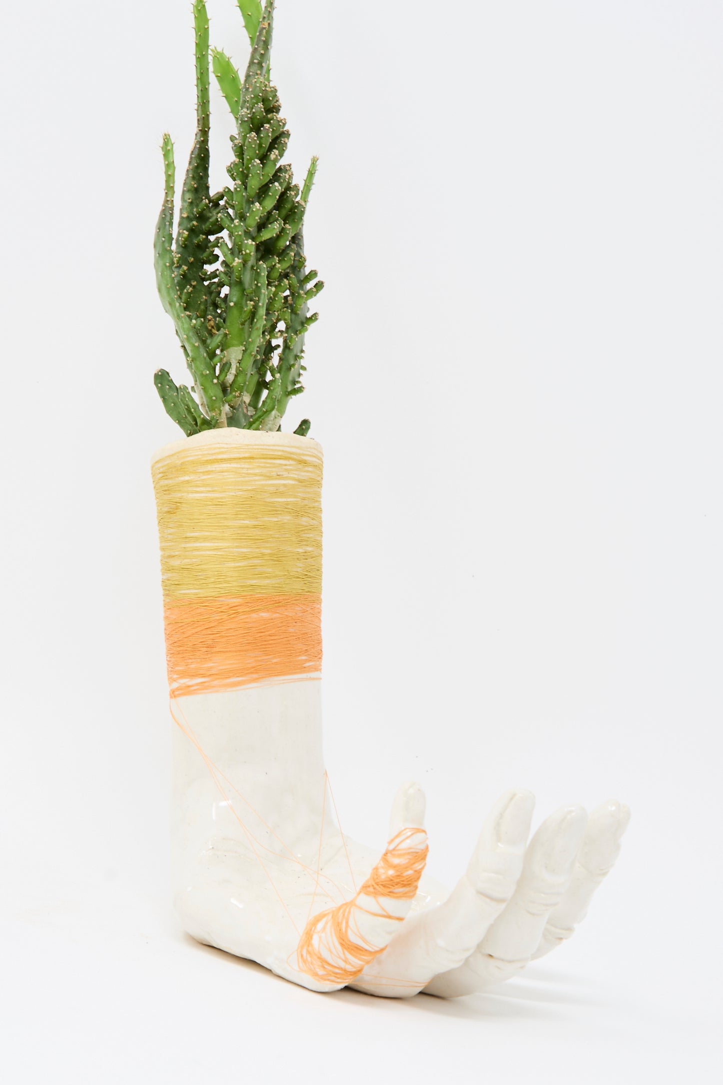 The Ceramic Hand Sculpture with Yellow and Orange Thread by Monty J features a green cactus plant in a handmade ceramic vessel, accented with yellow and orange thread around the wrist and fingers. Made in Brooklyn.