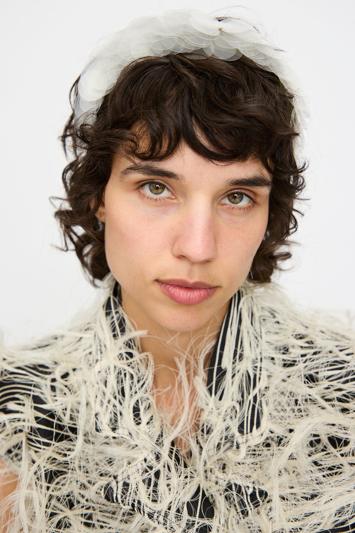 A person with short curly hair and light eyes looks at the camera, wearing a textured garment with feathery details and the Niccolò Pasqualetti Sfocatura Headband in Khaki against a plain background.