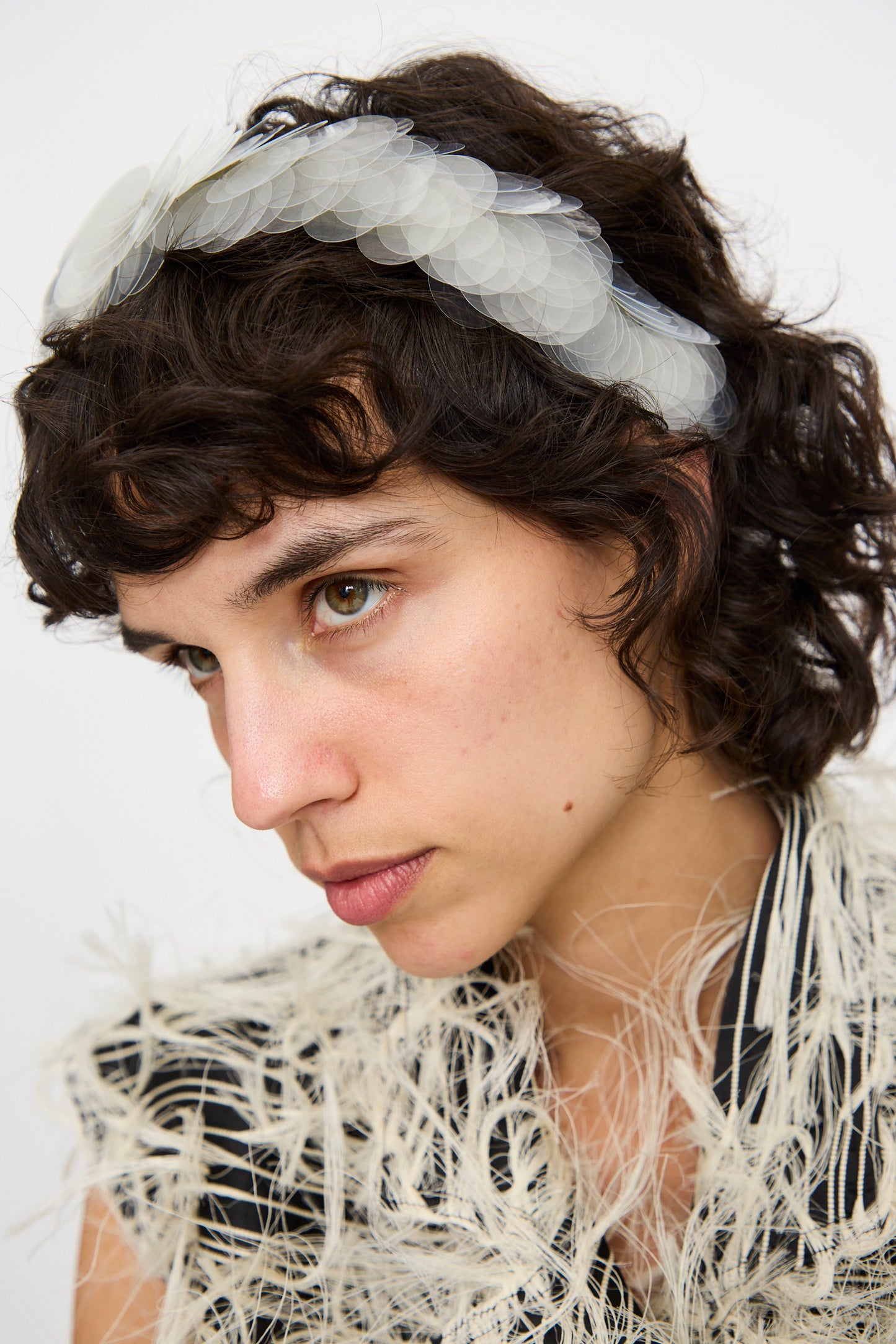 A person with short, wavy dark hair wears the Niccolò Pasqualetti Sfocatura Headband in Khaki and a textured, feather-like garment, looking slightly to the side.