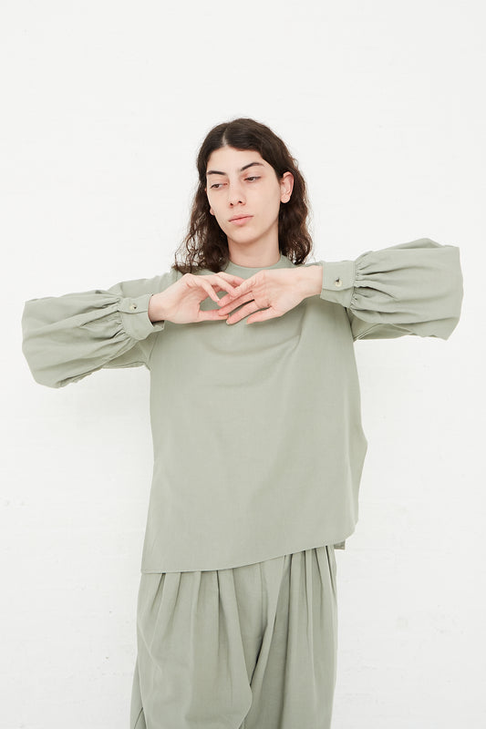 A woman in a Cotton Twill Puff Sleeve Blouse in Agave by Black Crane. Front view. Model's arms are raised to chest level.