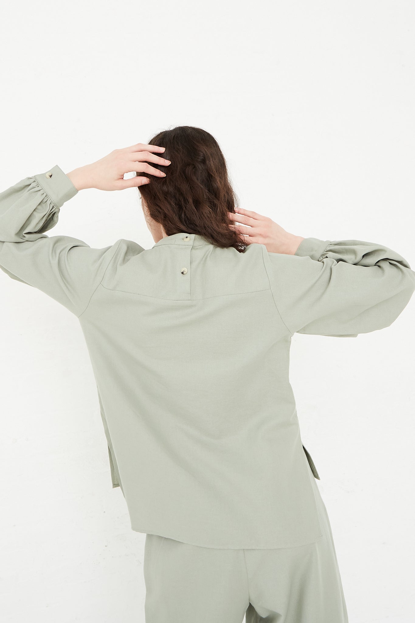 A woman in a Cotton Twill Puff Sleeve Blouse in Agave by Black Crane is standing in front of a white wall. Back view.