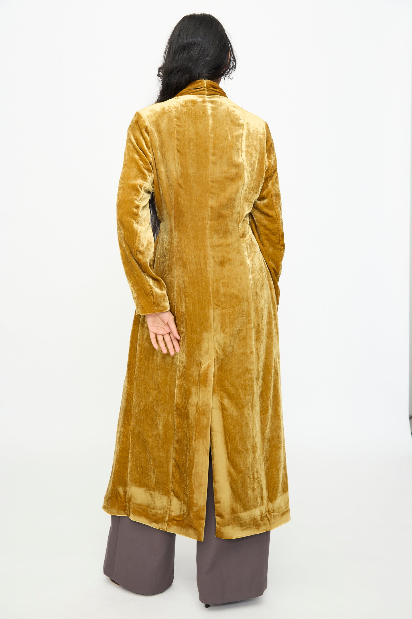 A person with long dark hair stands facing away, wearing the Theresa Coat in Amber by Olivia Cashmore—a luxurious gold viscose silk wrap—over brown pants, set against a plain white background.