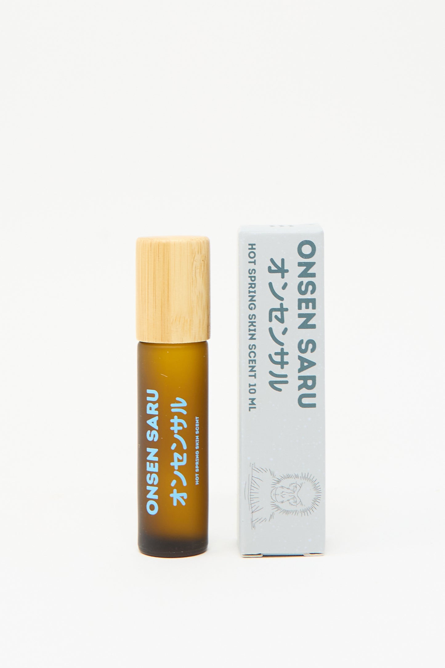 A brown glass bottle with a bamboo cap is placed next to a white box labeled "Onsen Saru Hot Spring Skin Scent 10 mL," adorned with Japanese text and an illustration of a monkey. This natural perfume embodies the essence of Japanese Matsu pine, delivering a refreshing unisex fragrance.