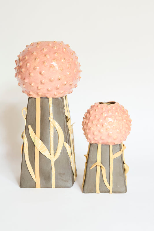 Two Pearce Williams Allium Vase Large pieces are shown: flower-inspired vases with textured round pink tops and tall rectangular gray bases, each featuring a raised yellow leaf design, displayed against a plain white background.
