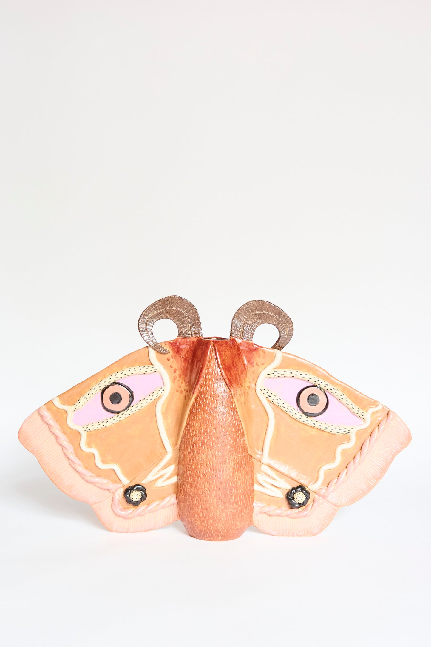 The Emperor Moth in Rust by Pearce Williams is a glazed stoneware sculpture featuring large orange and pink wings, black and white circular patterns, and textured brown antennae, displayed on a plain white background.