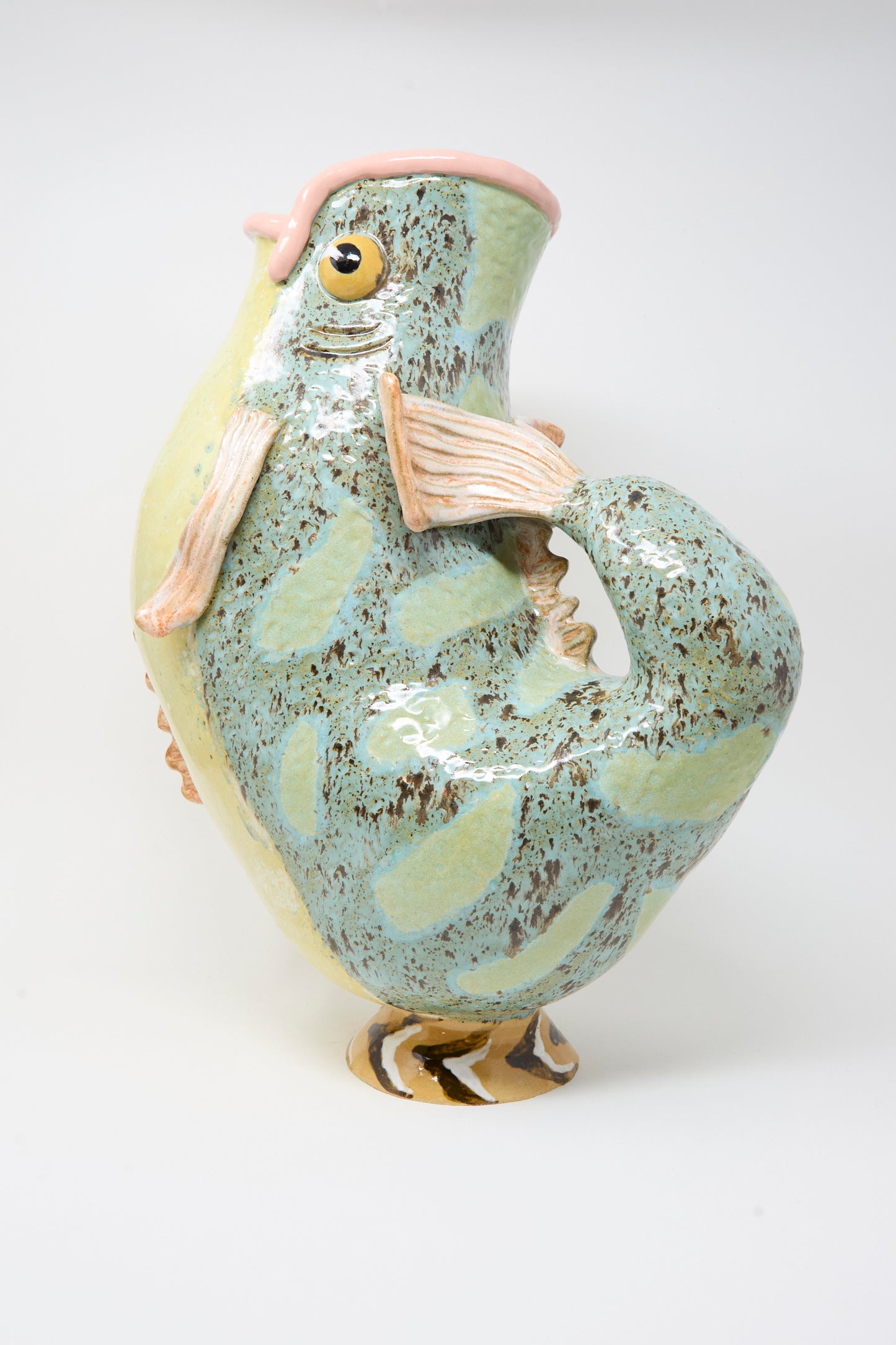 The Pearce Williams Fish Pitcher I is a colorful glazed vase shaped like a fish, featuring a mottled green and yellow finish with pink details and an open mouth as the vase opening.