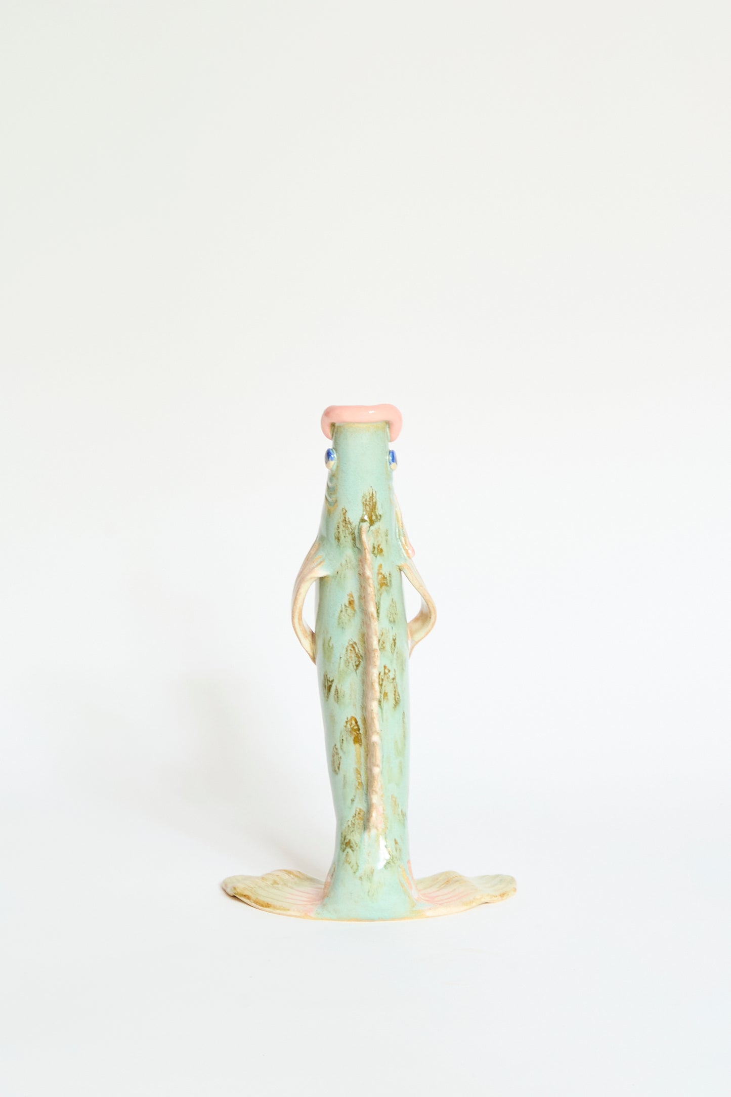 The Pearce Williams Fish Vase Large in Green is a handmade, upright, fish-shaped glazed stoneware vase with a light green and beige finish, blue eyes, and a pink mouth, shown against a plain white background.