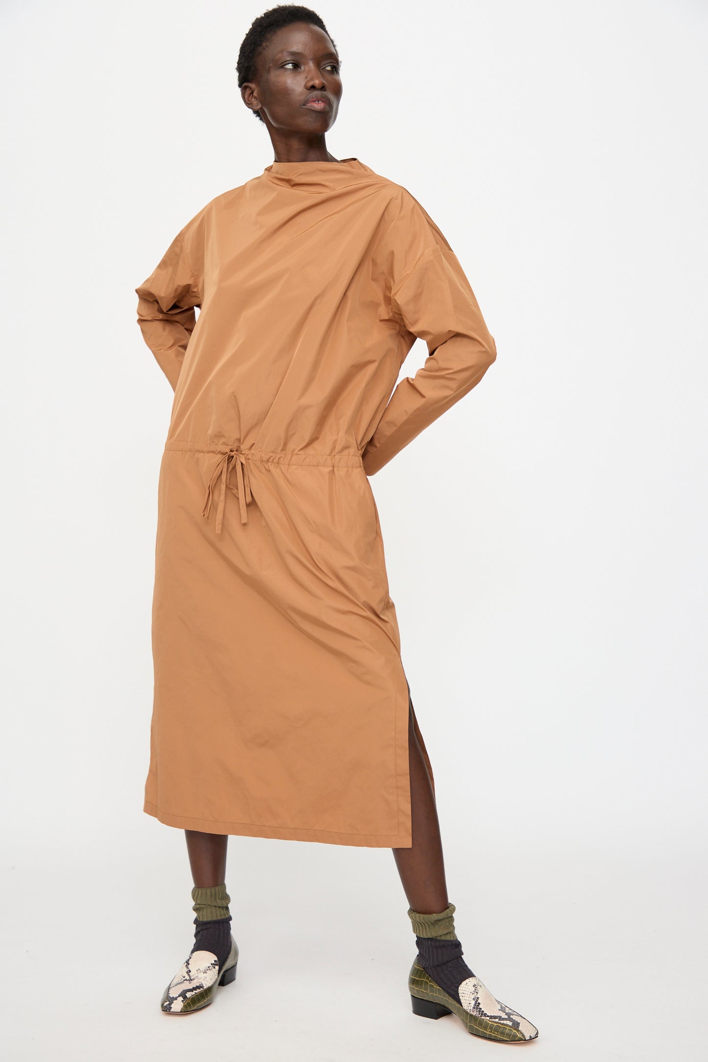 A person wears the Rachel Comey Alberry Dress in Copper—a loose, long sleeve, water-resistant taffeta midi with a drawstring waist and side slit—paired with green socks and snakeskin shoes, against a plain white background.