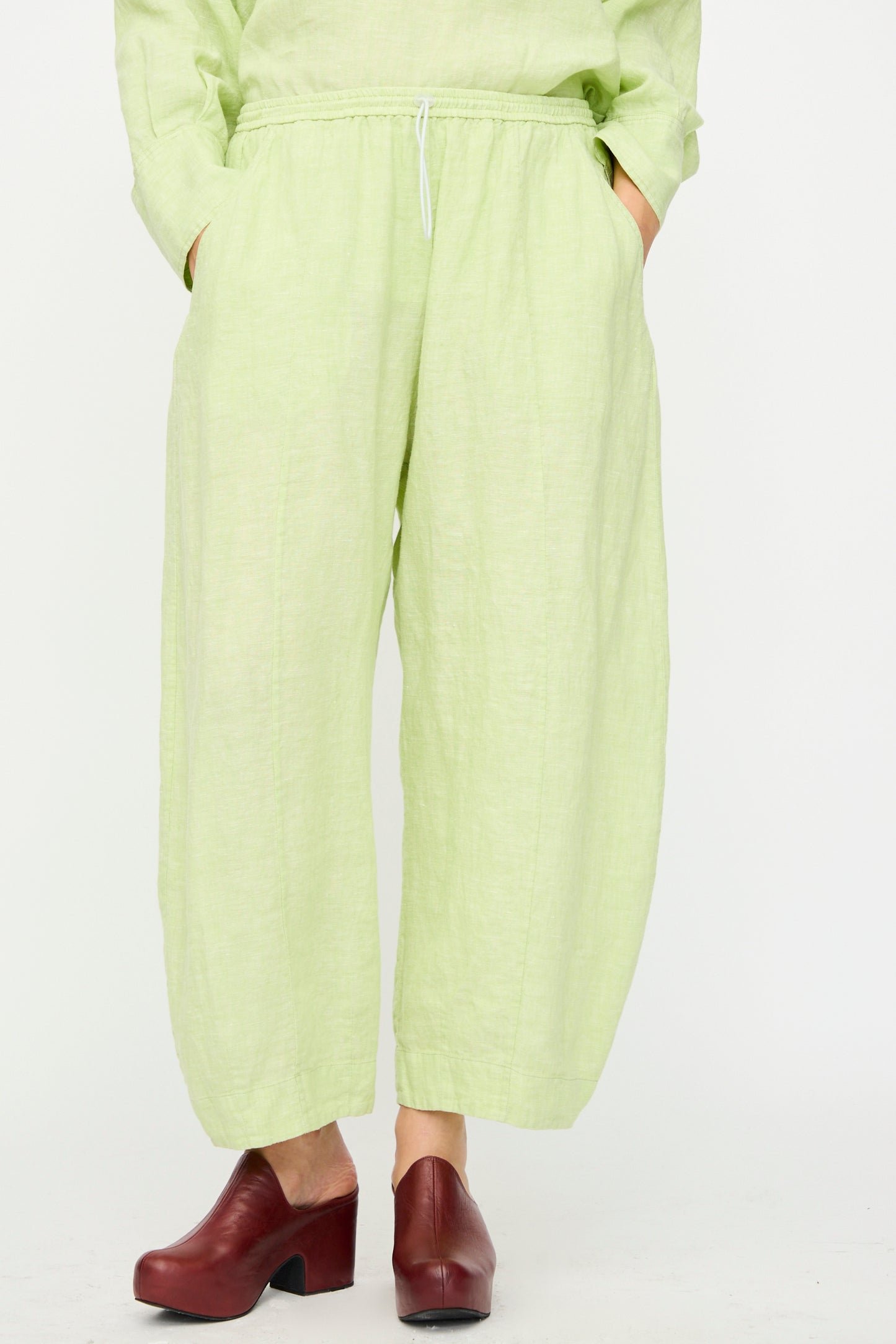 Model wears the Rachel Comey Ashbolt Pant in White Sage—loose, wide-leg textured pants—with hands in pockets and red clogs, standing against a plain white background.