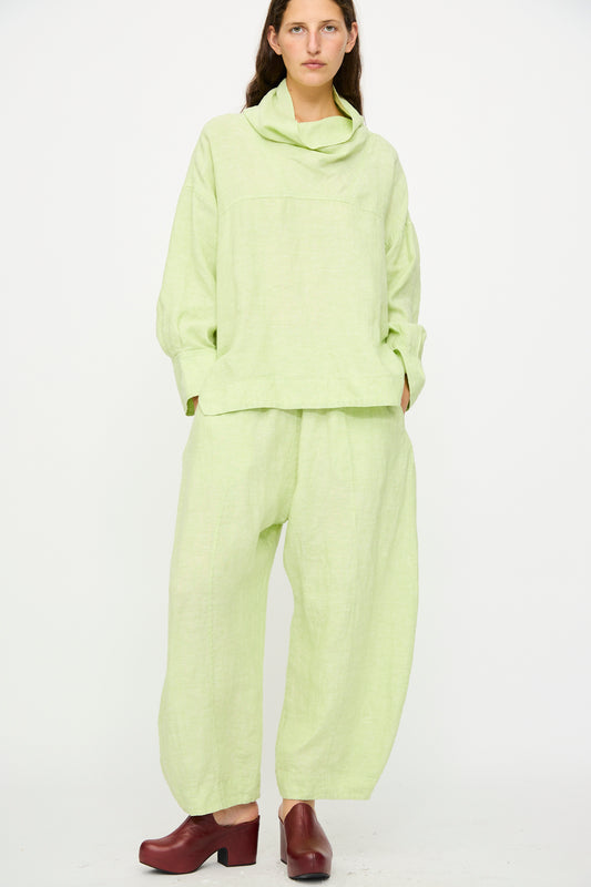 A person wears the Rachel Comey Ashbolt Pant in White Sage—a loose-fitting, wide-leg pant with an elastic waistband—paired with a matching linen top and brown clogs, posing against a plain white background.