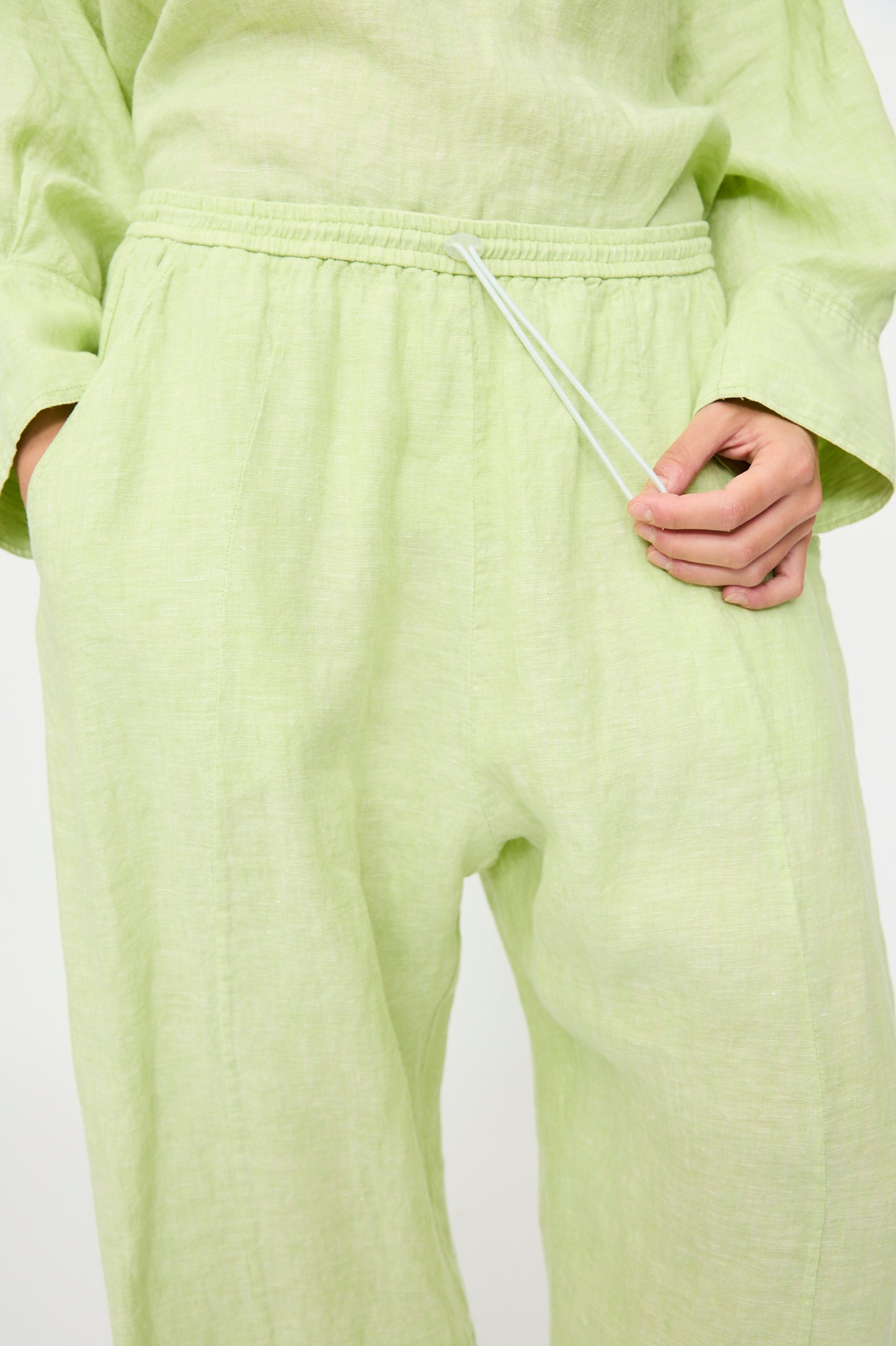 A person wears Rachel Comey's Ashbolt Pant in White Sage, a light green textured wide-leg style with an elastic waistband and drawstring, shown waist down with one hand in a pocket and the other holding the drawstring.