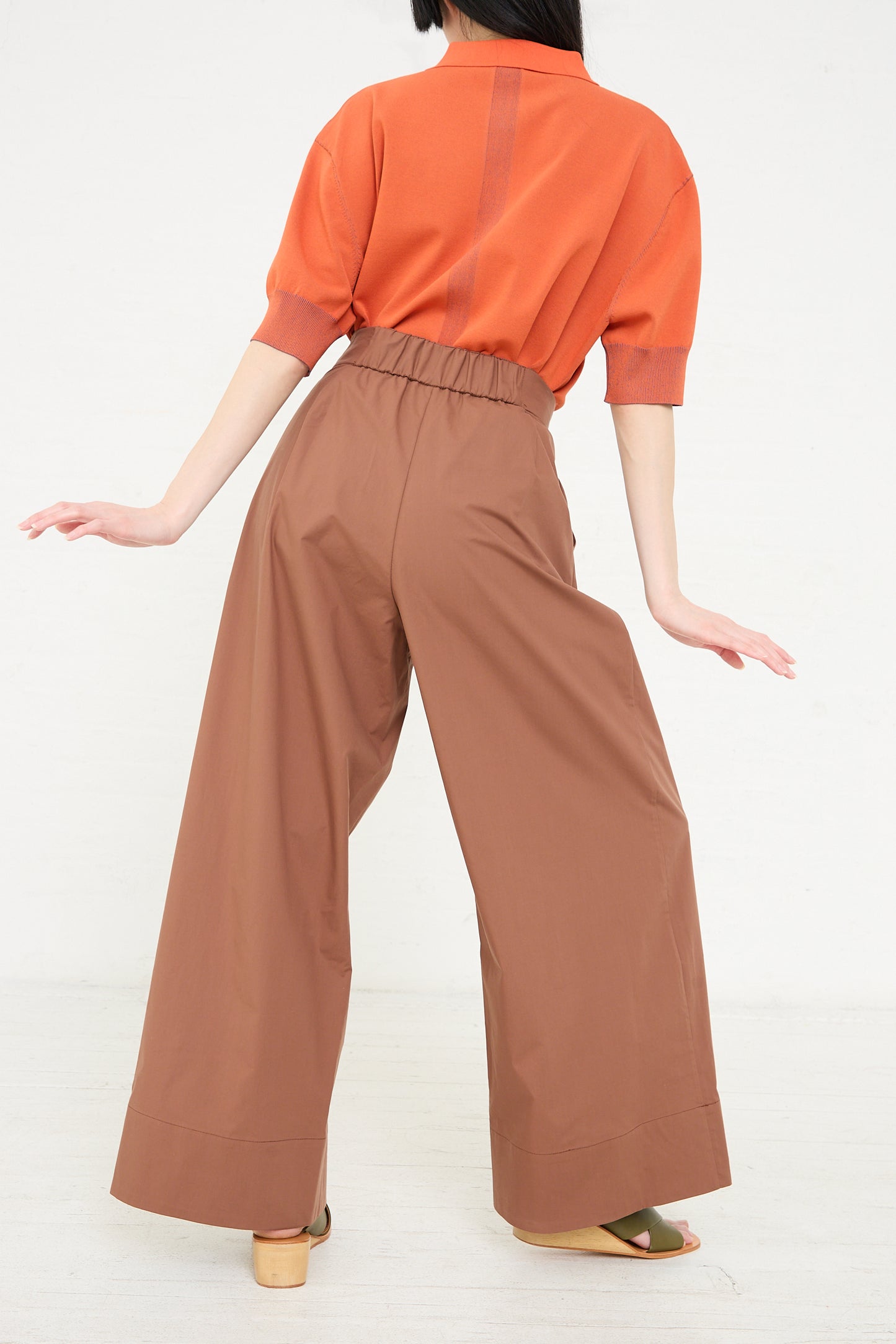 A woman is shown from the back wearing Rachel Comey's Coxsone Pant in Sienna, a pair of brown wide-leg, high waisted trousers and an orange top, posed with her hands lifted slightly away from her body.
