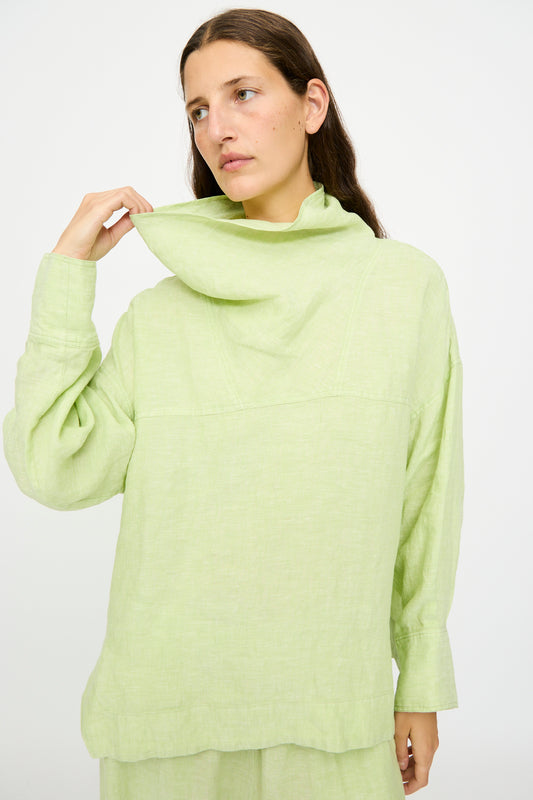 A woman wearing the Rachel Comey Fiamma Top in White Sage, a relaxed fit linen long-sleeve, holds up the collar with one hand while looking off to the side against a plain white background.