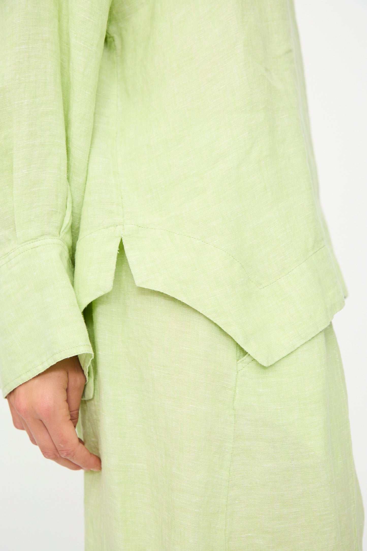 A close-up of a person wearing the Rachel Comey Fiamma Top in White Sage, highlighting the shirt cuff, side slit, and hem details of this relaxed-fit, lightweight linen ensemble.
