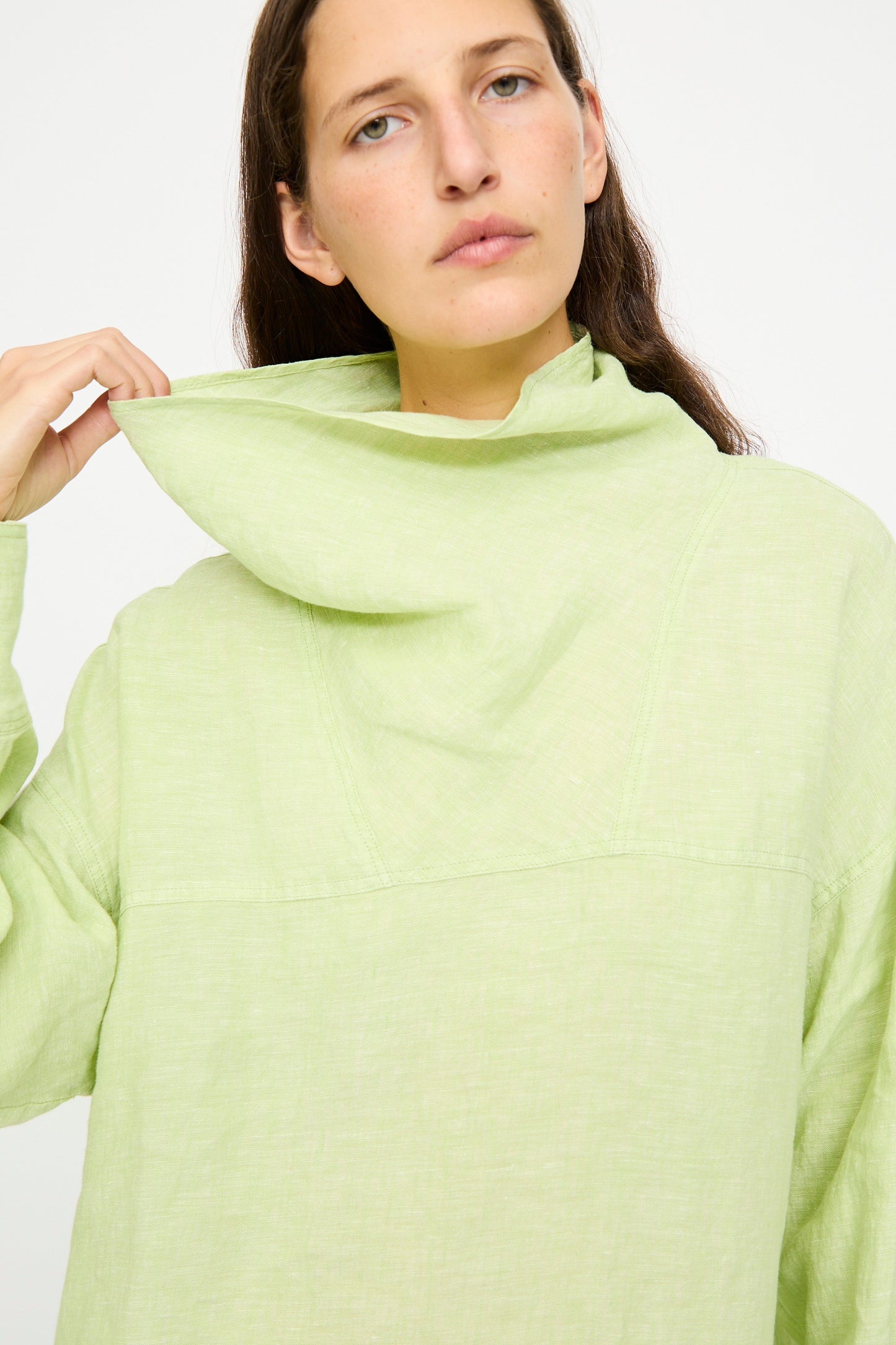 A person wearing the Rachel Comey Fiamma Top in White Sage—a lightweight linen, loose high-collar long-sleeve—holds the collar with one hand against a plain white background.