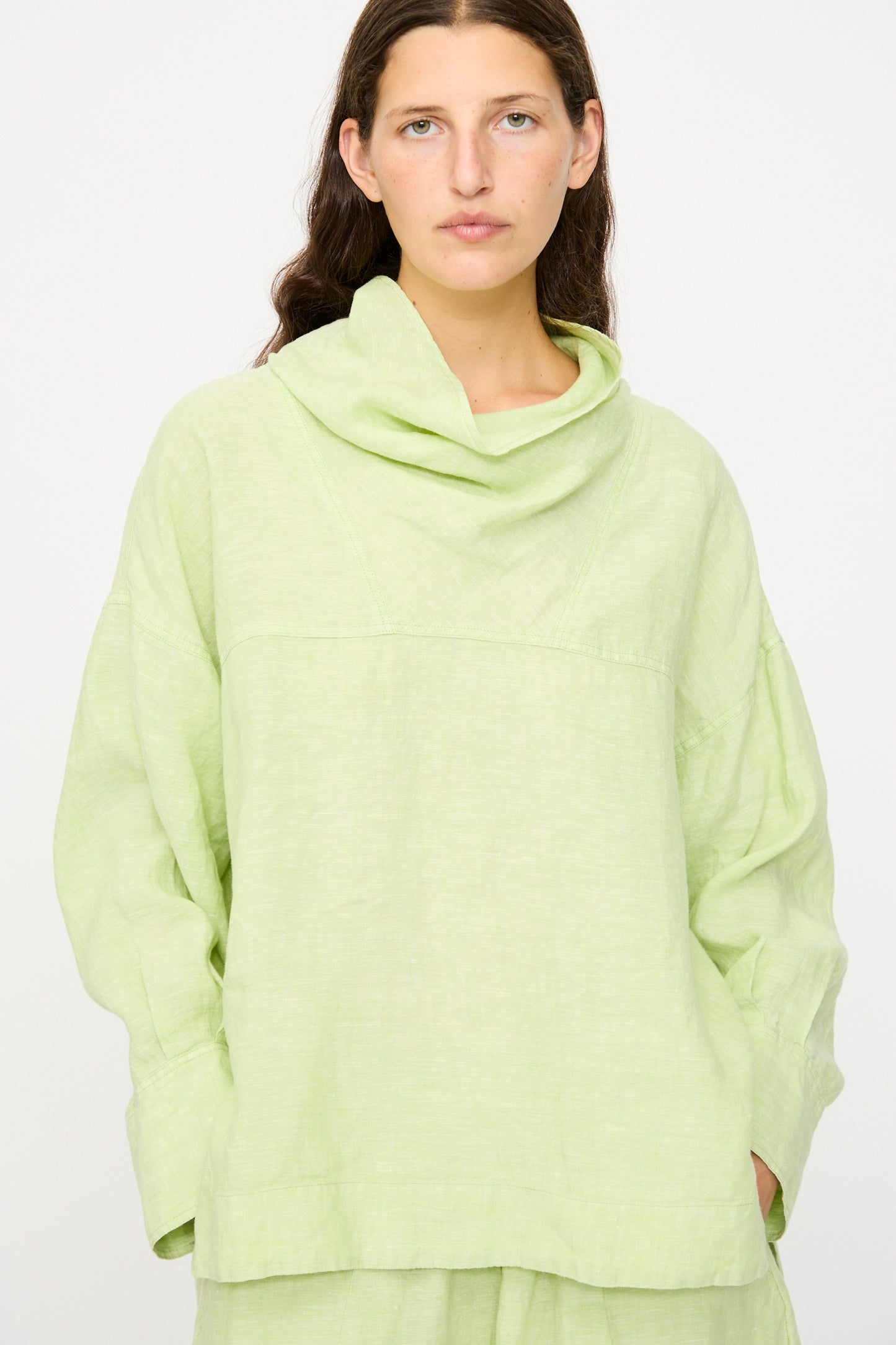 A person with long brown hair wears the Rachel Comey Fiamma Top in White Sage, a lightweight linen, long-sleeve top with a wide cowl neck and relaxed fit, standing against a plain white background.