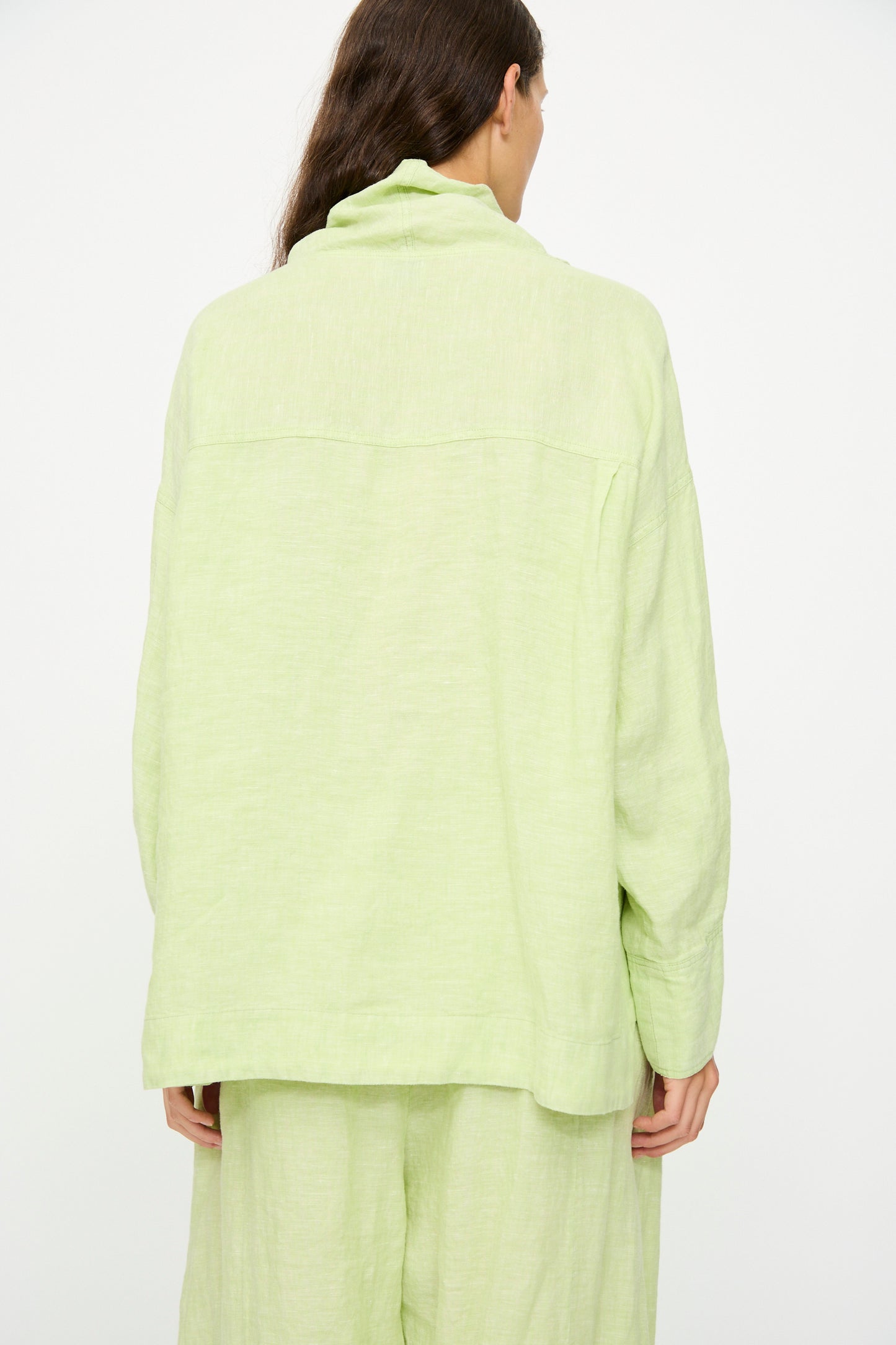 A person with long brown hair, shown from the back, wears the Rachel Comey Fiamma Top in White Sage— a light green, relaxed-fit linen long-sleeve top—paired with matching pants, against a plain white background.