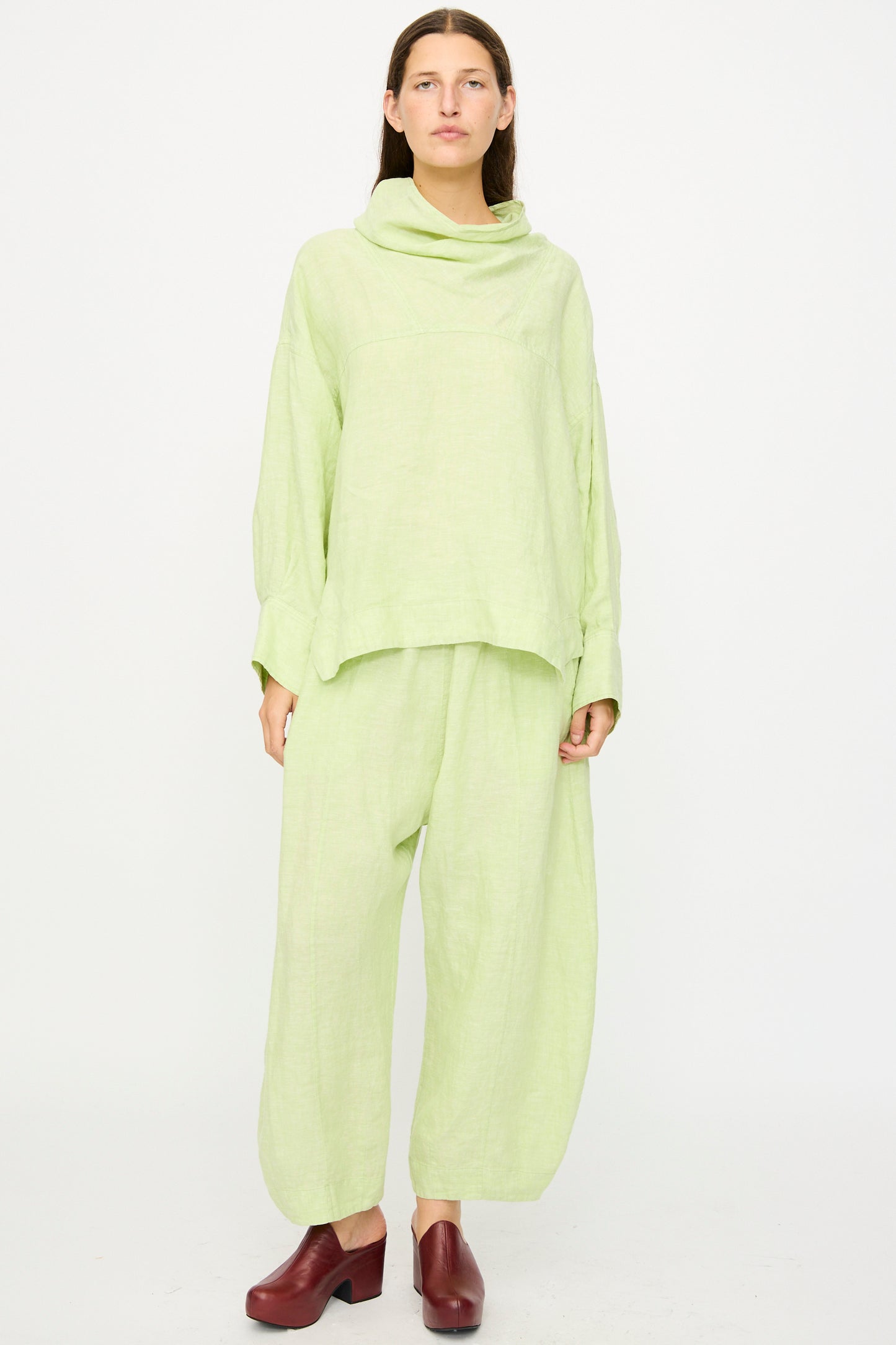 A person stands against a white background wearing the Rachel Comey Fiamma Top in White Sage, a relaxed fit, lightweight linen long-sleeve top with matching wide-leg pants and dark red shoes.