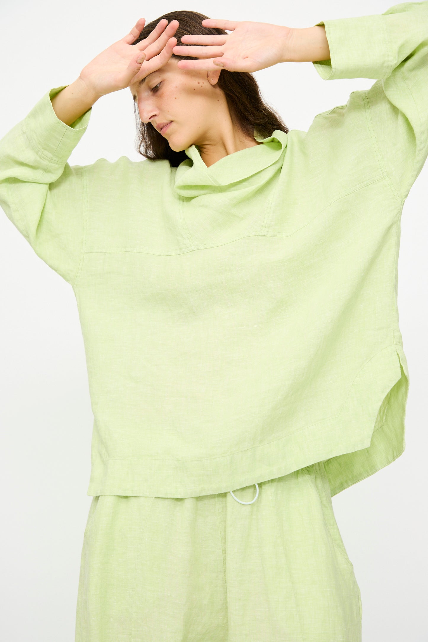 Wearing the Rachel Comey Fiamma Top in White Sage, a person stands with their hands raised near their face against a plain background.