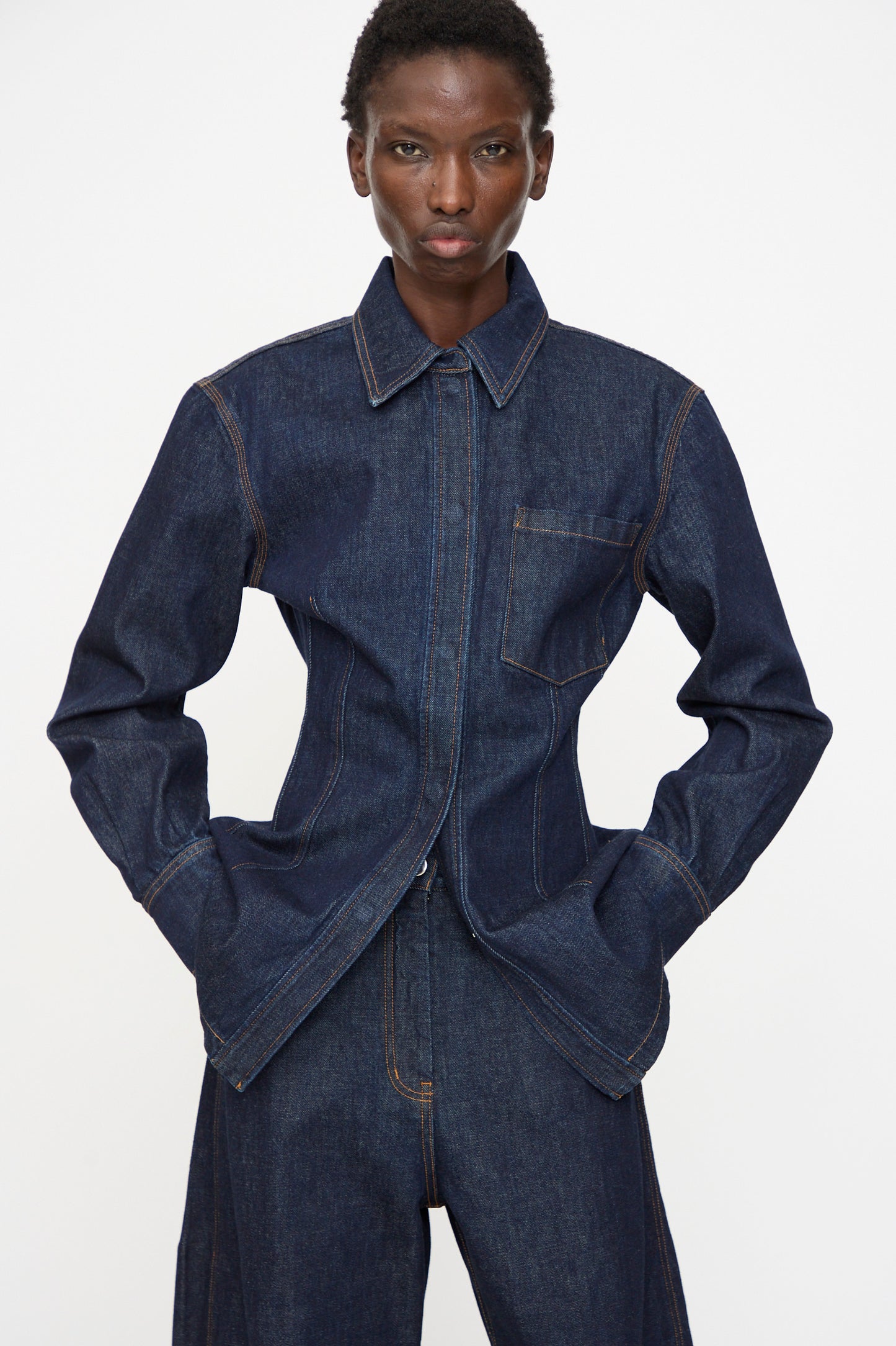 A person wears the Rachel Comey Filt Top in Raw Wash—a dark denim shirt—layered under a matching jacket and pants with tan stitching, standing against a plain white background with hands in pockets.
