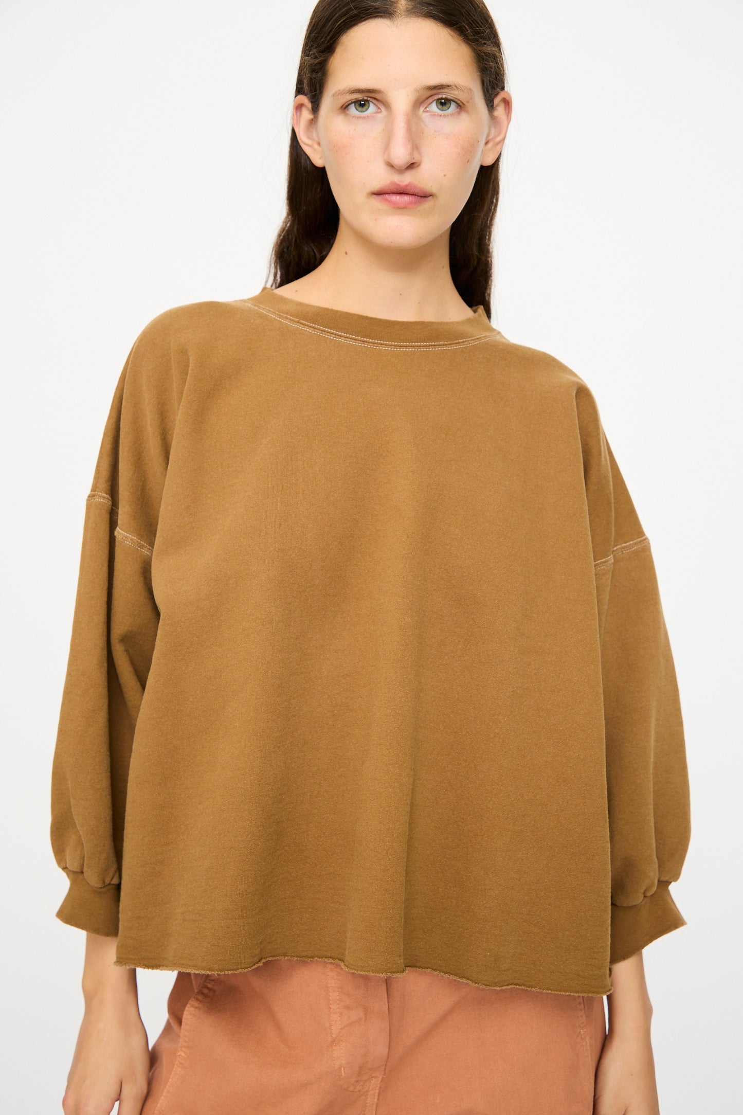 A woman with long brown hair models the Rachel Comey Fond Sweatshirt in Bronze—an oversized, genderless piece—paired with peach pants against a plain white background.