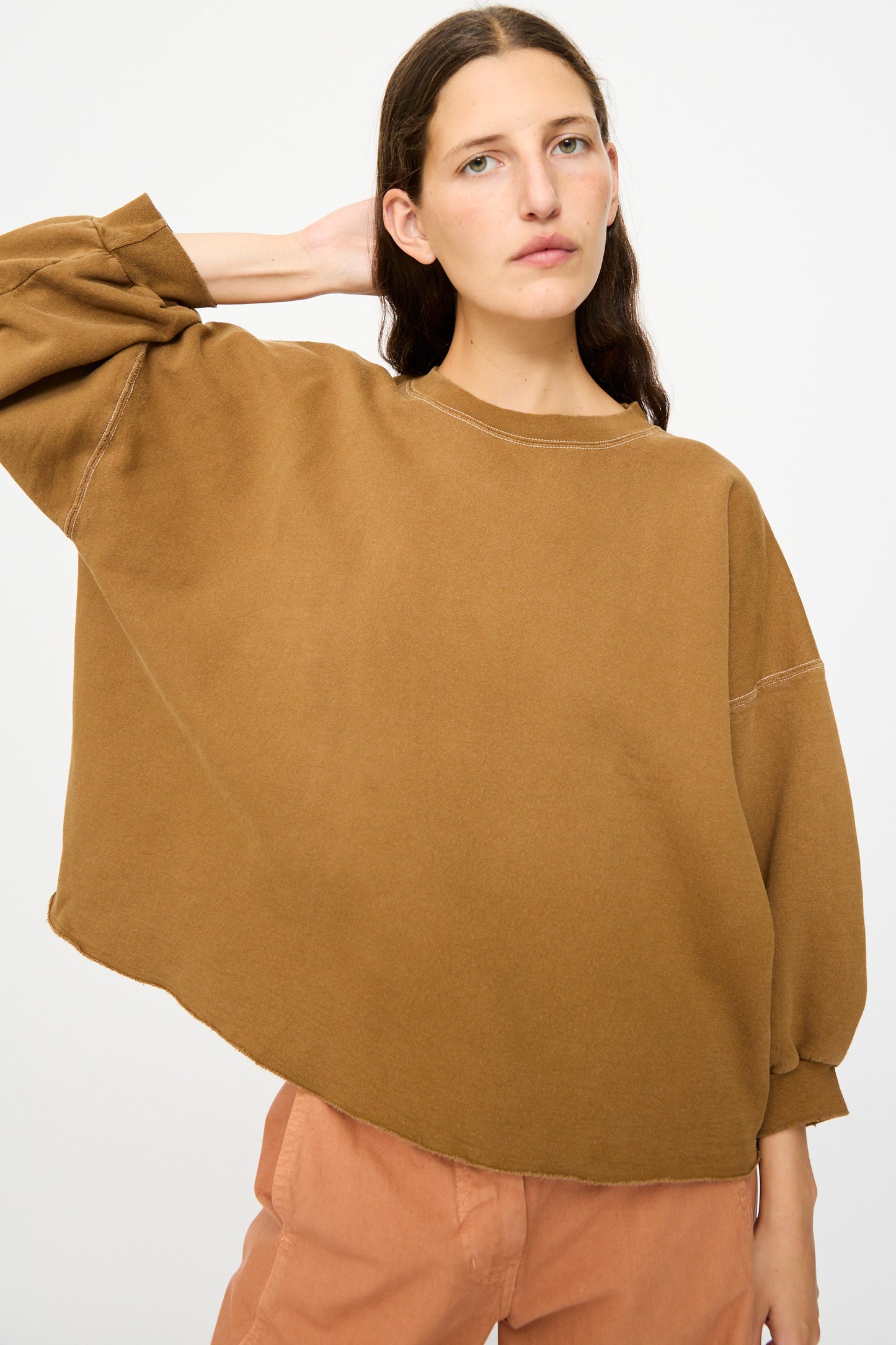 A person with long brown hair wears the Rachel Comey Fond Sweatshirt in Bronze and peach pants, standing against a plain light background with one hand behind their head.