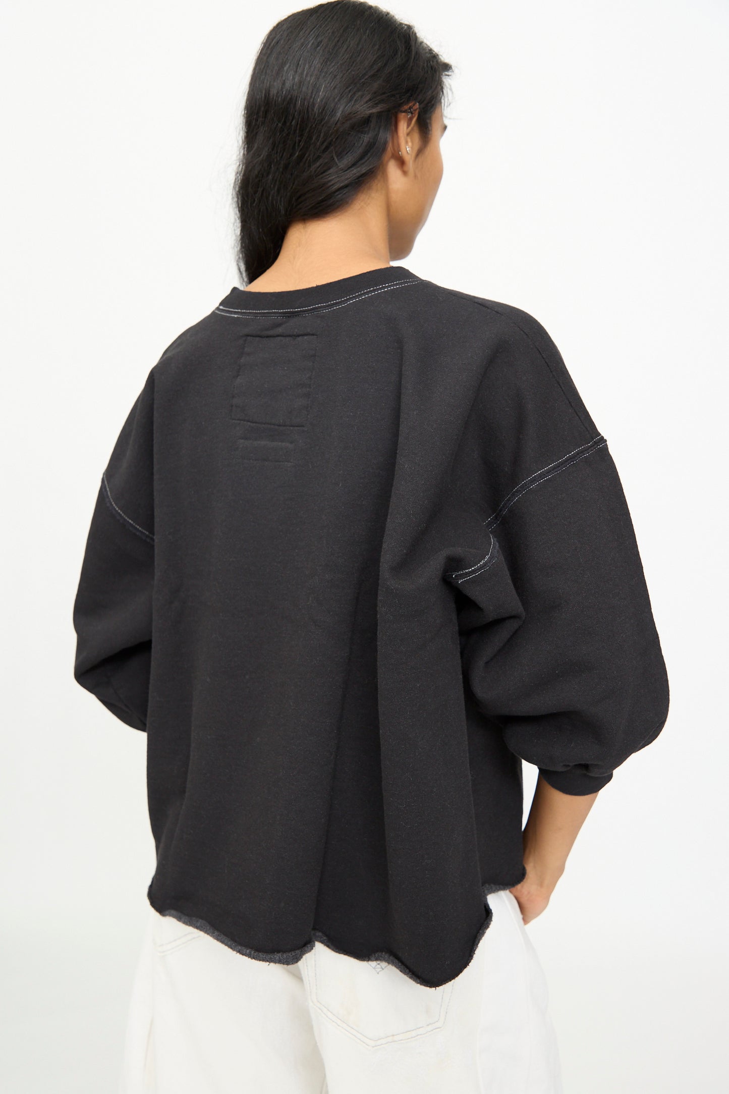 A person with long dark hair is shown from the back against a plain white background, wearing the Rachel Comey Fond Sweatshirt in Charcoal—an oversized black sweatshirt featuring visible white stitching and a raw edge hemline—paired with off-white pants.