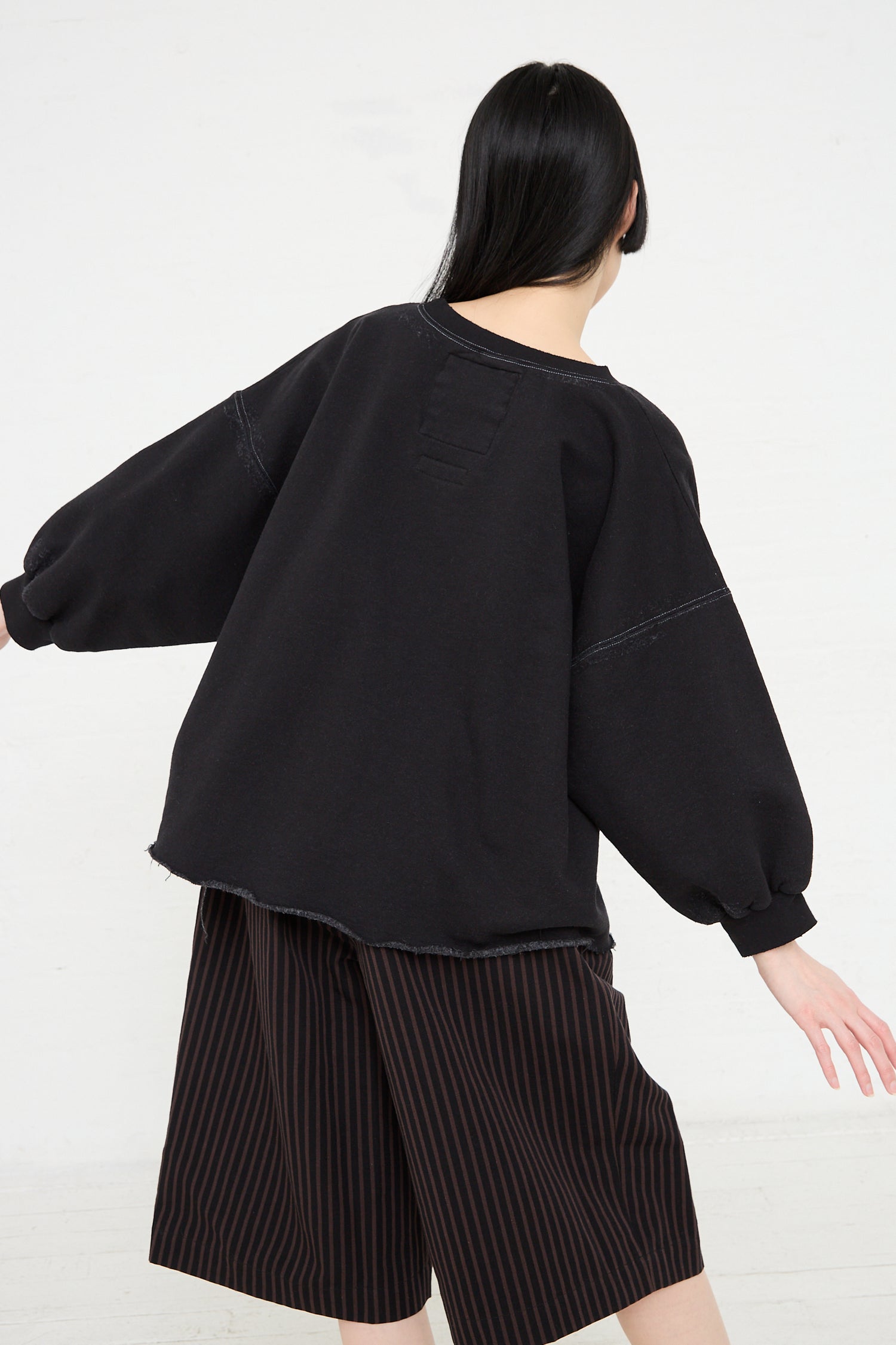 A person from behind wearing a Rachel Comey Fond Sweatshirt in Charcoal, a genderless oversized sweatshirt made of black cotton blend, and striped shorts.