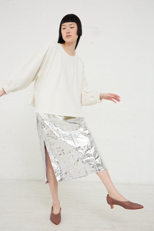 A woman posing in a casual Rachel Comey Fond Sweatshirt in Dirty White paired with a metallic silver skirt and brown heeled loafers.