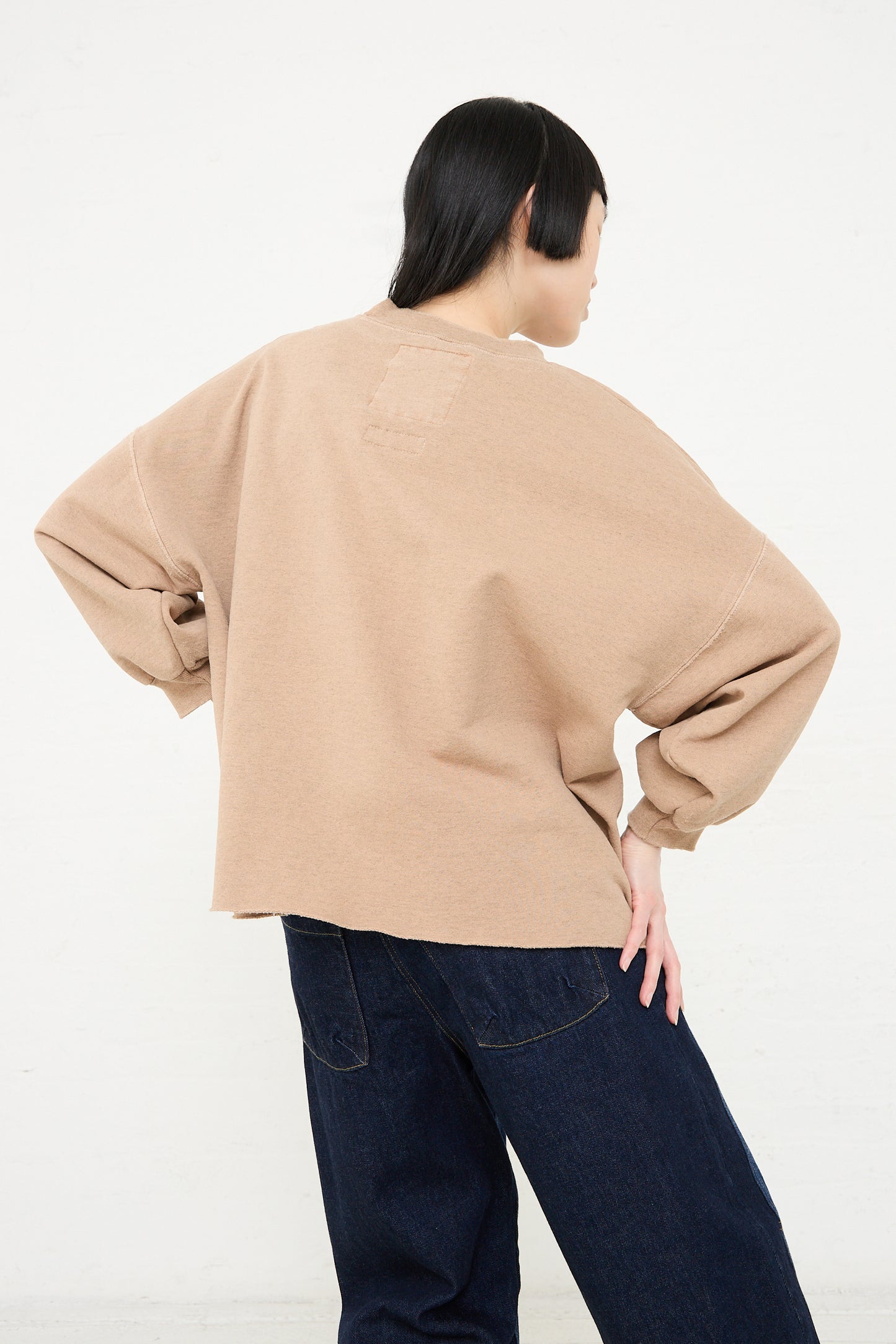 Person in oversized, distressed Rachel Comey Fond Sweatshirt in Hazelnut and blue jeans standing with back to the camera against a white background.