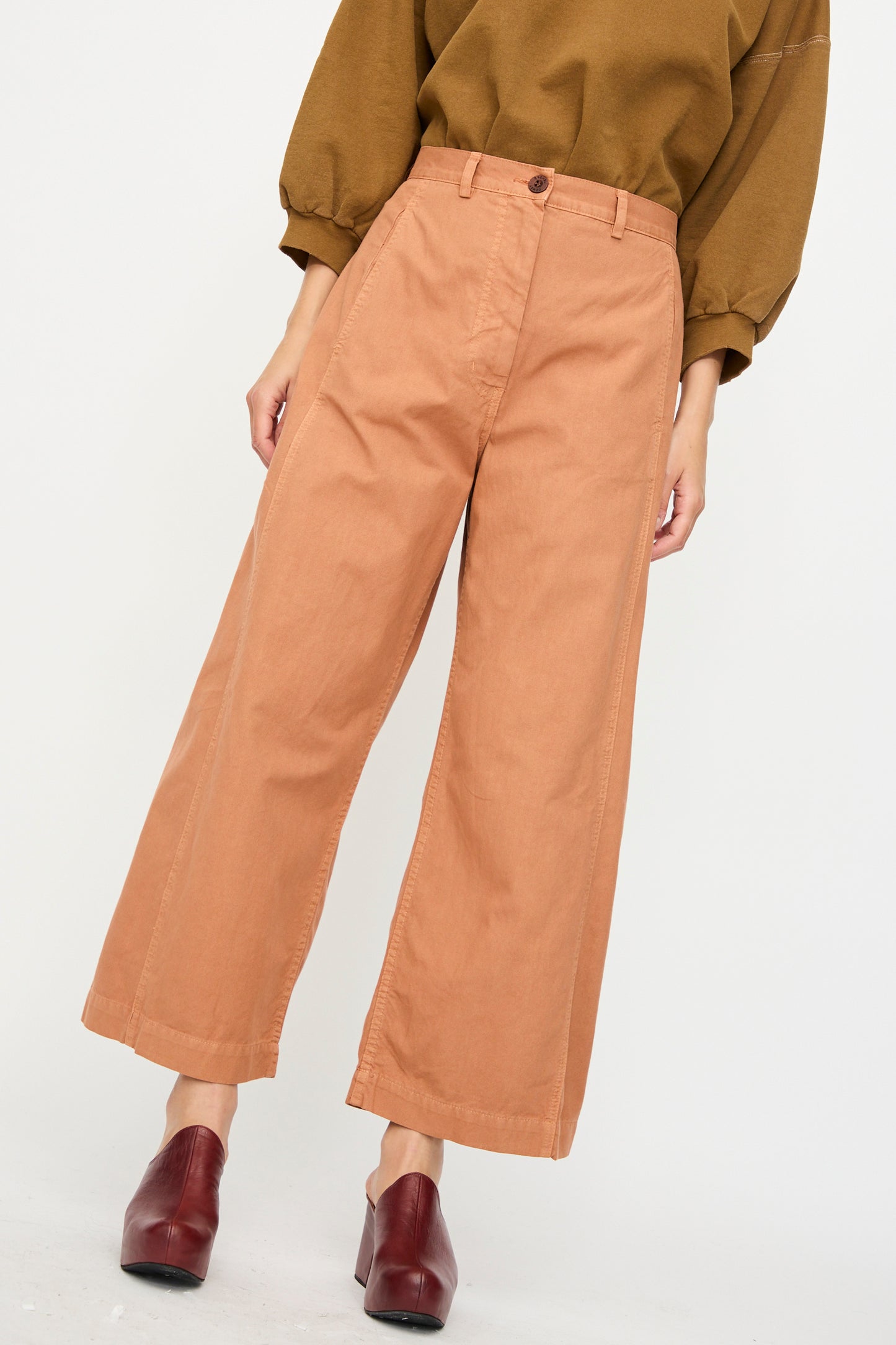Person wears the Rachel Comey Garra Pant in Copper paired with a brown long-sleeve top and dark brown heeled shoes, standing against a plain light background.