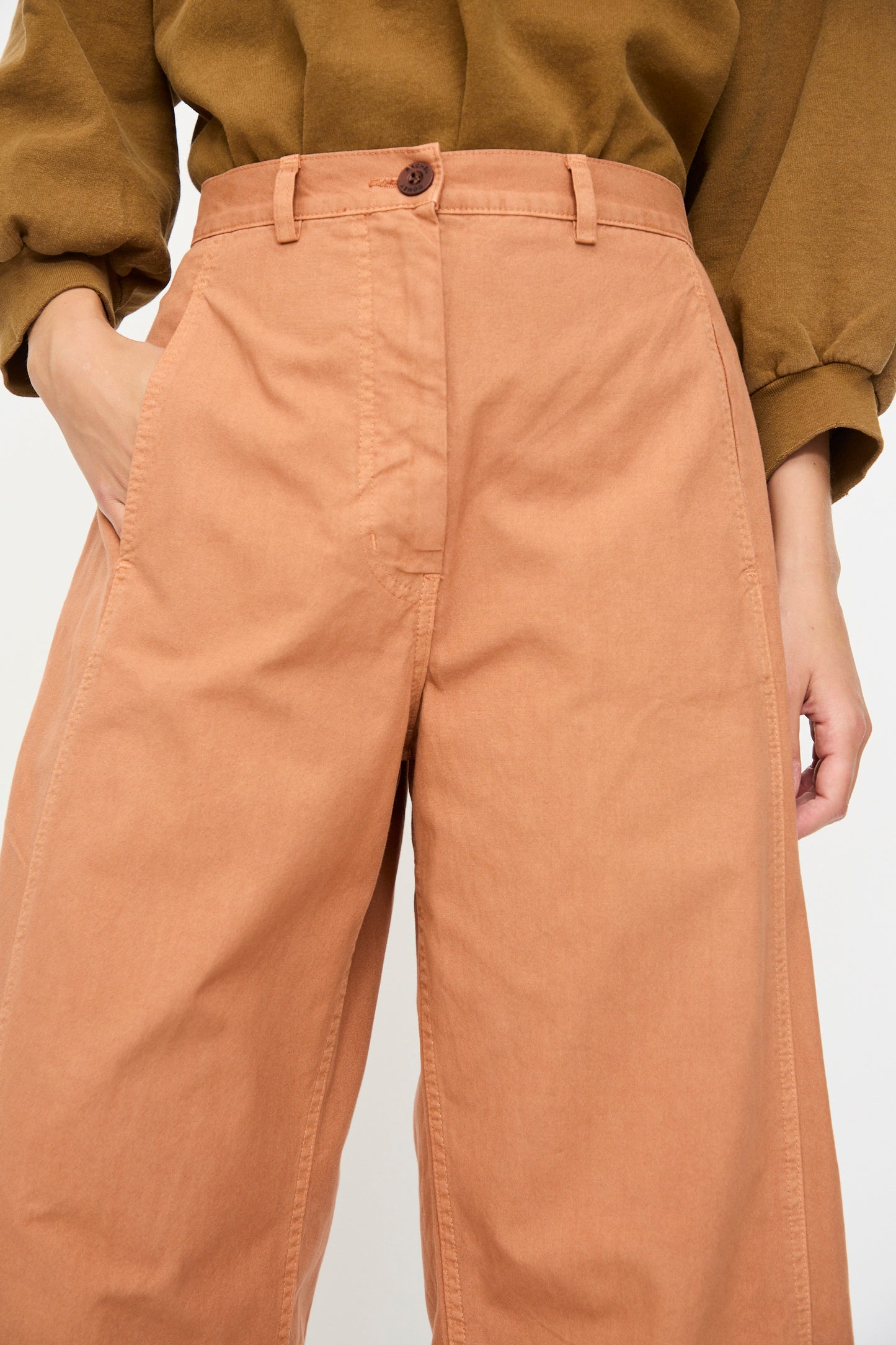 Model wearing the Rachel Comey Garra Pant in Copper, styled as high-waisted wide-leg pants, paired with an olive green long-sleeve top, one hand in pocket; visible from waist to mid-thigh.