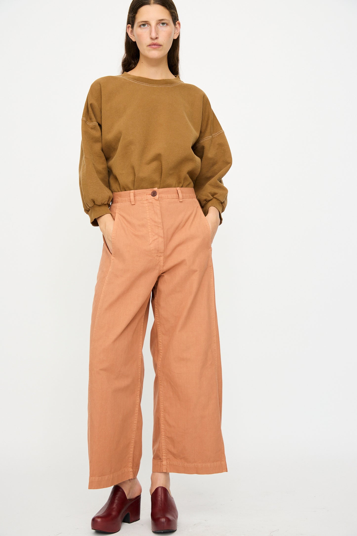 A person stands against a plain background wearing a loose brown sweatshirt, Rachel Comey Garra Pant in Copper, and dark red shoes.