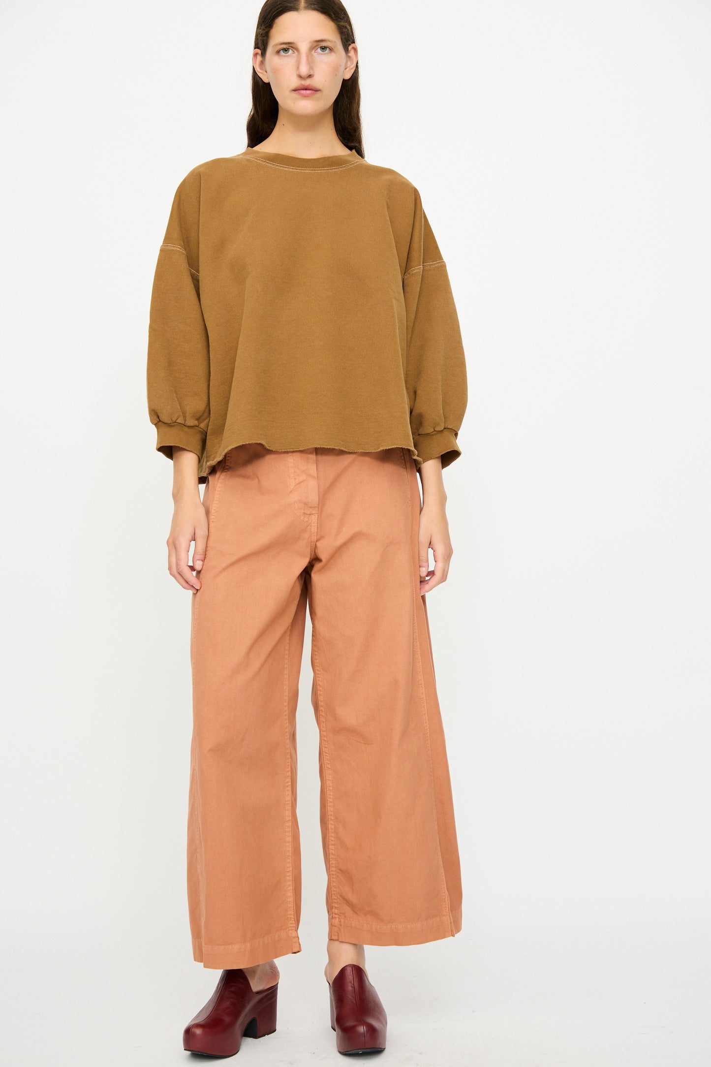 A person stands against a white background wearing a loose brown sweater, Rachel Comey's Garra Pant in Copper, and maroon heeled shoes.