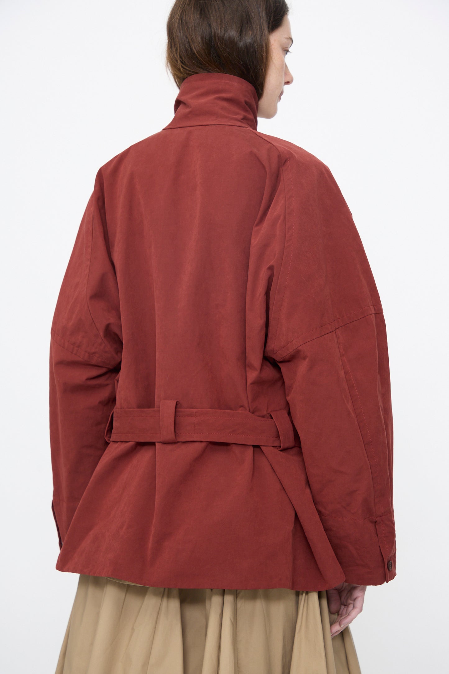 A person shown from the back wears the Rachel Comey Gosland Trench in Garnet, a relaxed-fit, belted jacket with a high collar, styled over a beige skirt.