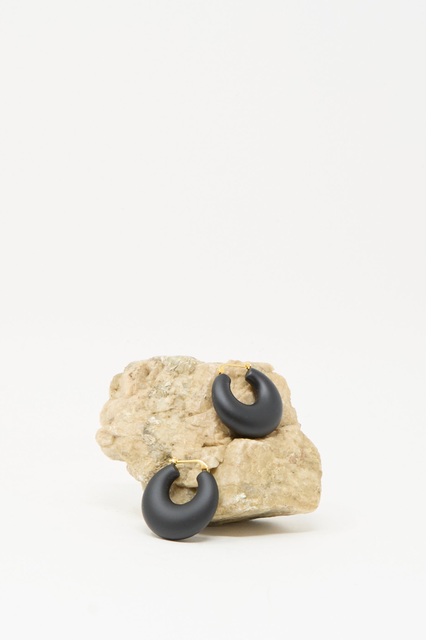 The Rachel Comey Grass Earrings in Black Matte are chunky acrylic hoops with gold hinged clasps, shown on a beige rock against a plain white background.