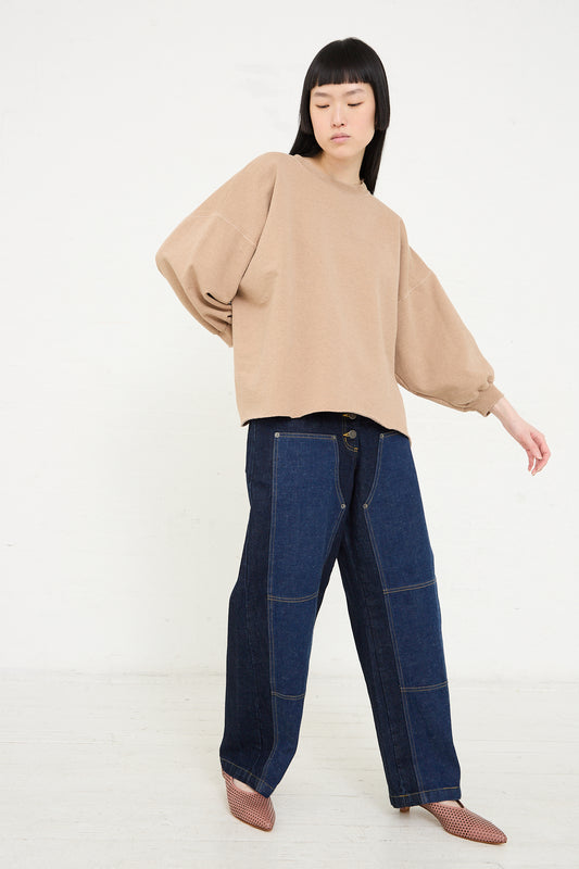 A person wearing an oversized beige top, Rachel Comey Japanese indigo denim carpenter pant, and checkered shoes stands with one hand on hip against a white background.
