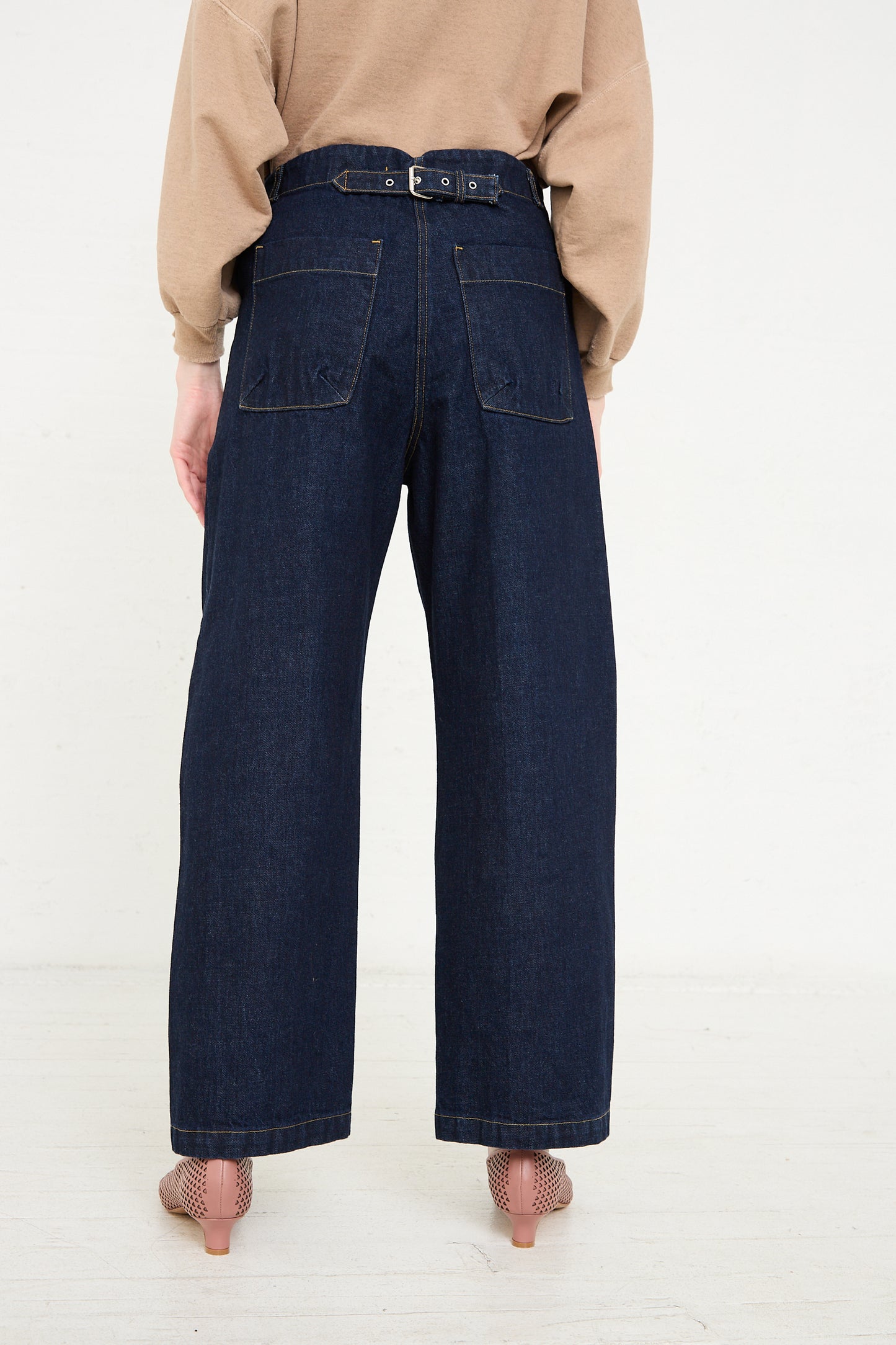 A rear view of a person wearing Rachel Comey's Handy Pant in Dark Indigo, paired with a brown belt, beige top, and mesh heels.
