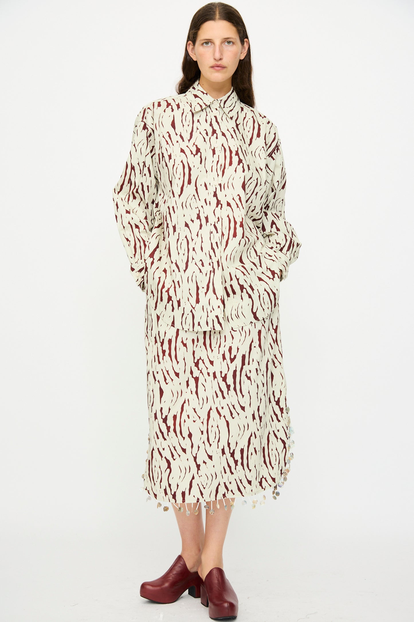 A person stands against a white background wearing the Rachel Comey Lastica Skirt in Cream with an abstract print, paired with a loose cream and burgundy shirt and burgundy platform shoes.