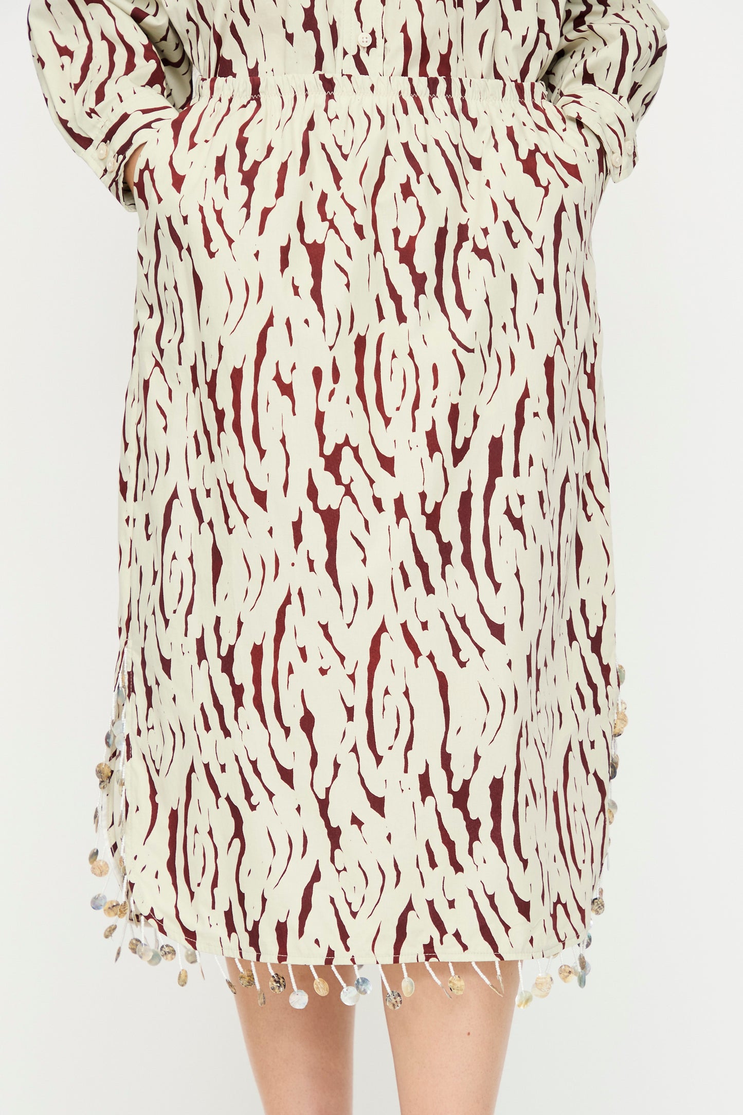 A person wearing the Rachel Comey Lastica Skirt in Cream—a cotton skirt with cream and maroon print, bead detailing on the hem, and pockets—shown from waist to knee.