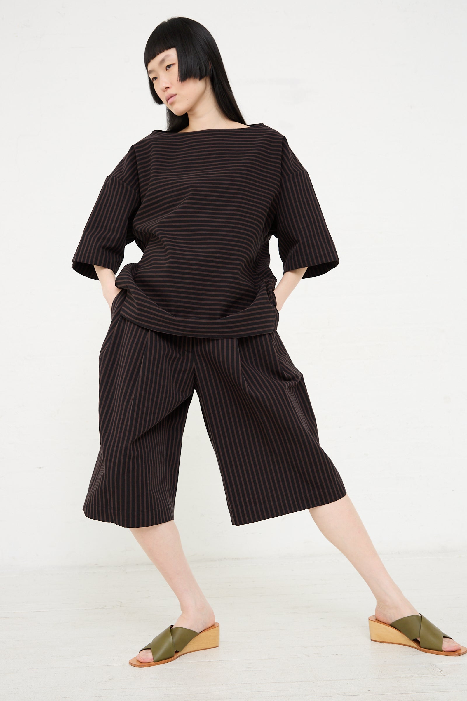 A woman posing in a Melio Short in Black and brown striped Bermuda top outfit made from a cotton/linen blend, paired with olive green sandals by Rachel Comey.