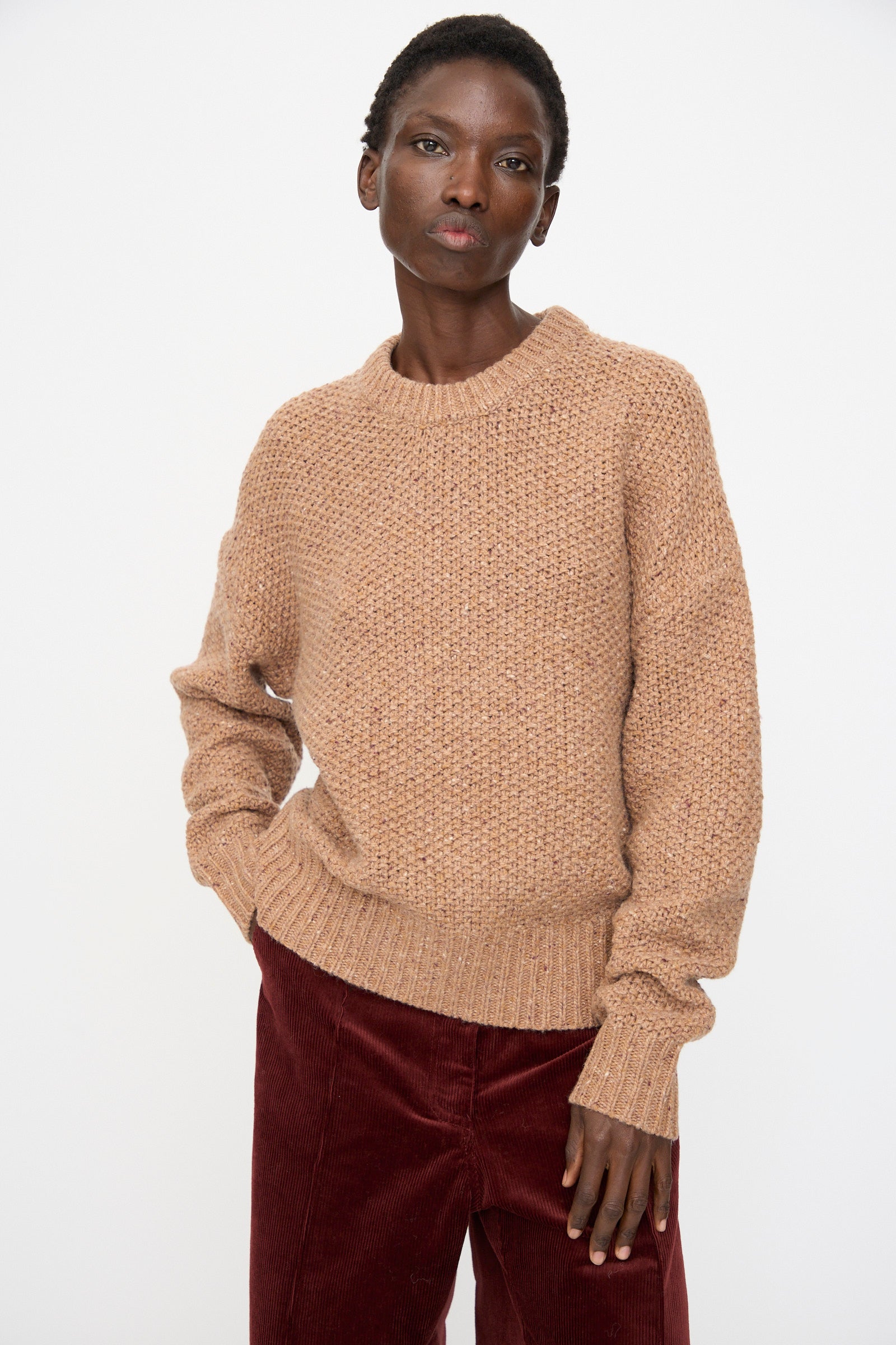 A person wears the Rachel Comey Prescot Sweater in Caramel and red corduroy pants while standing against a plain white background.