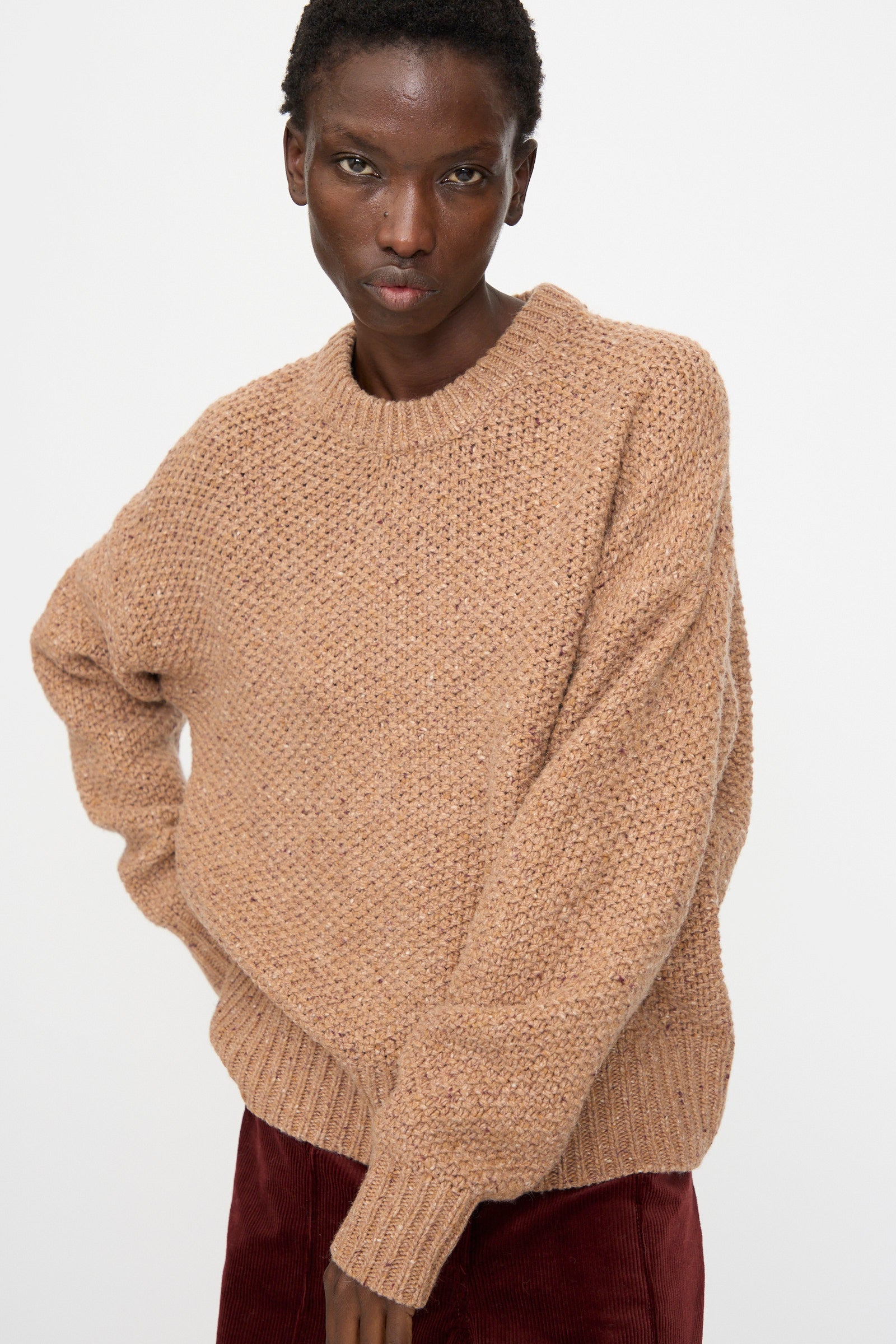 A person stands against a plain white background, looking directly at the camera, wearing the Rachel Comey Prescot Sweater in Caramel and maroon pants.