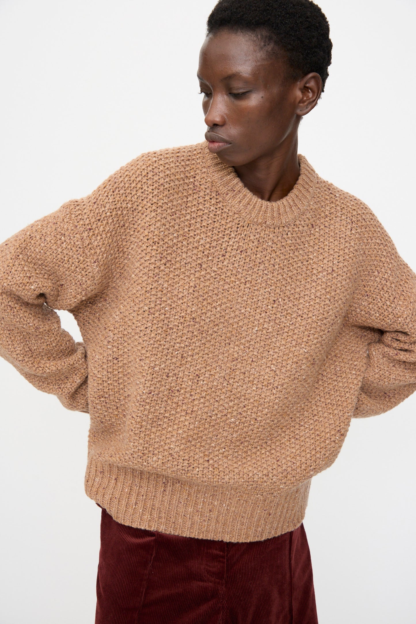 A person wears the Rachel Comey Prescot Sweater in Caramel with a burgundy skirt, standing against a plain white background and looking slightly to the side.