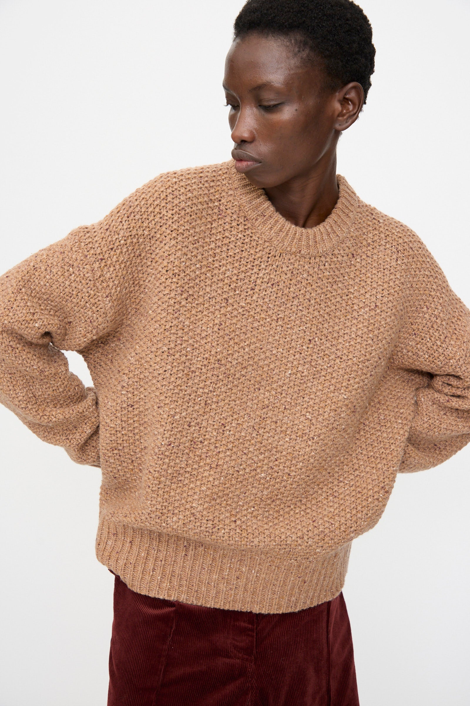 A person wears the Rachel Comey Prescot Sweater in Caramel with a burgundy skirt, standing against a plain white background and looking slightly to the side.