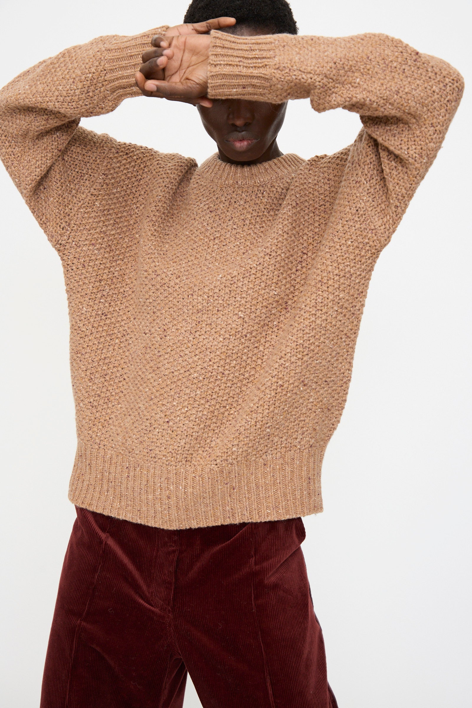 A person wearing the Rachel Comey Prescot Sweater in Caramel and burgundy corduroy pants stands with arms raised, partially covering their face.